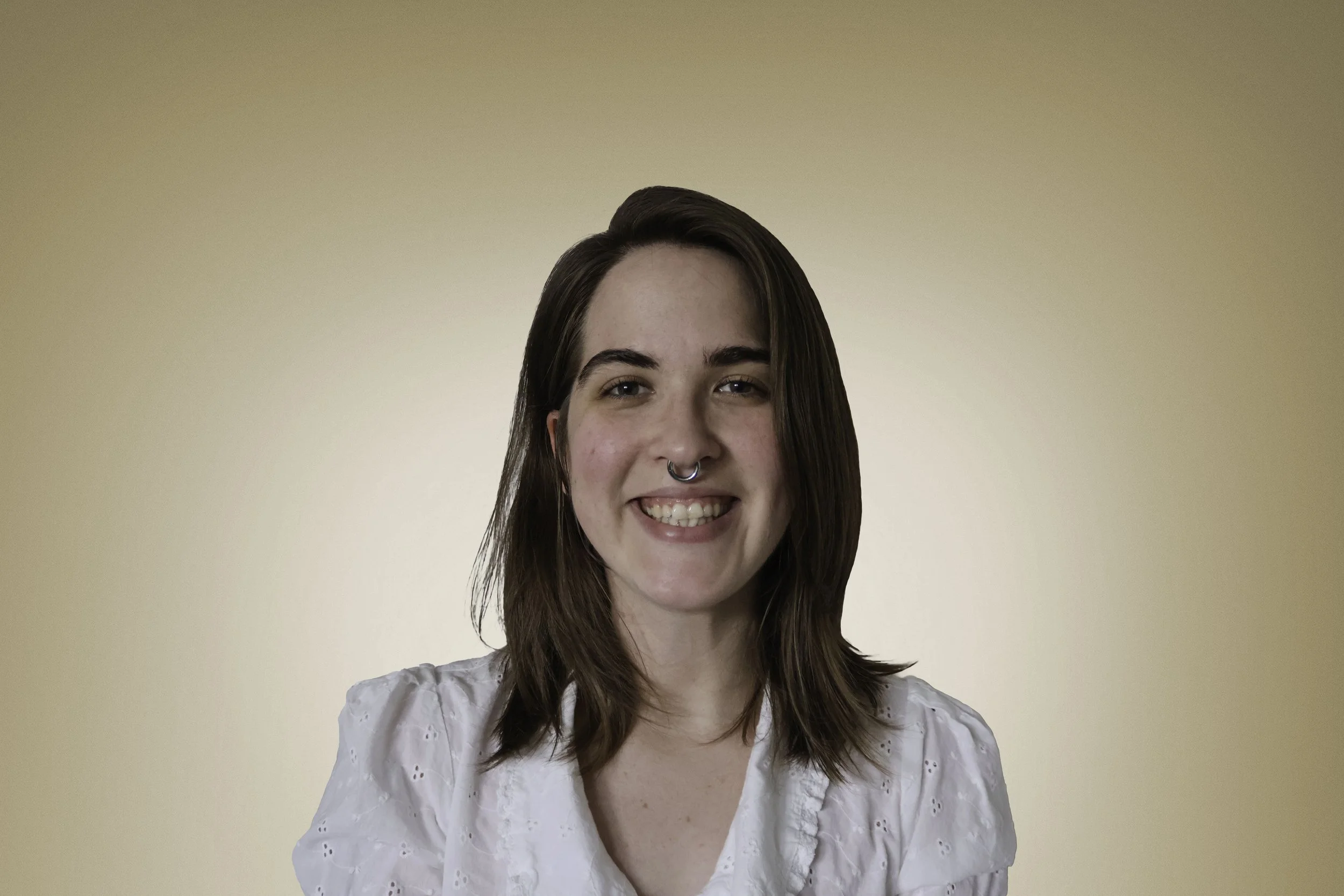 Portrait of a young woman with brown hair, smiling, wearing a light-colored blouse, standing against a plain beige background.