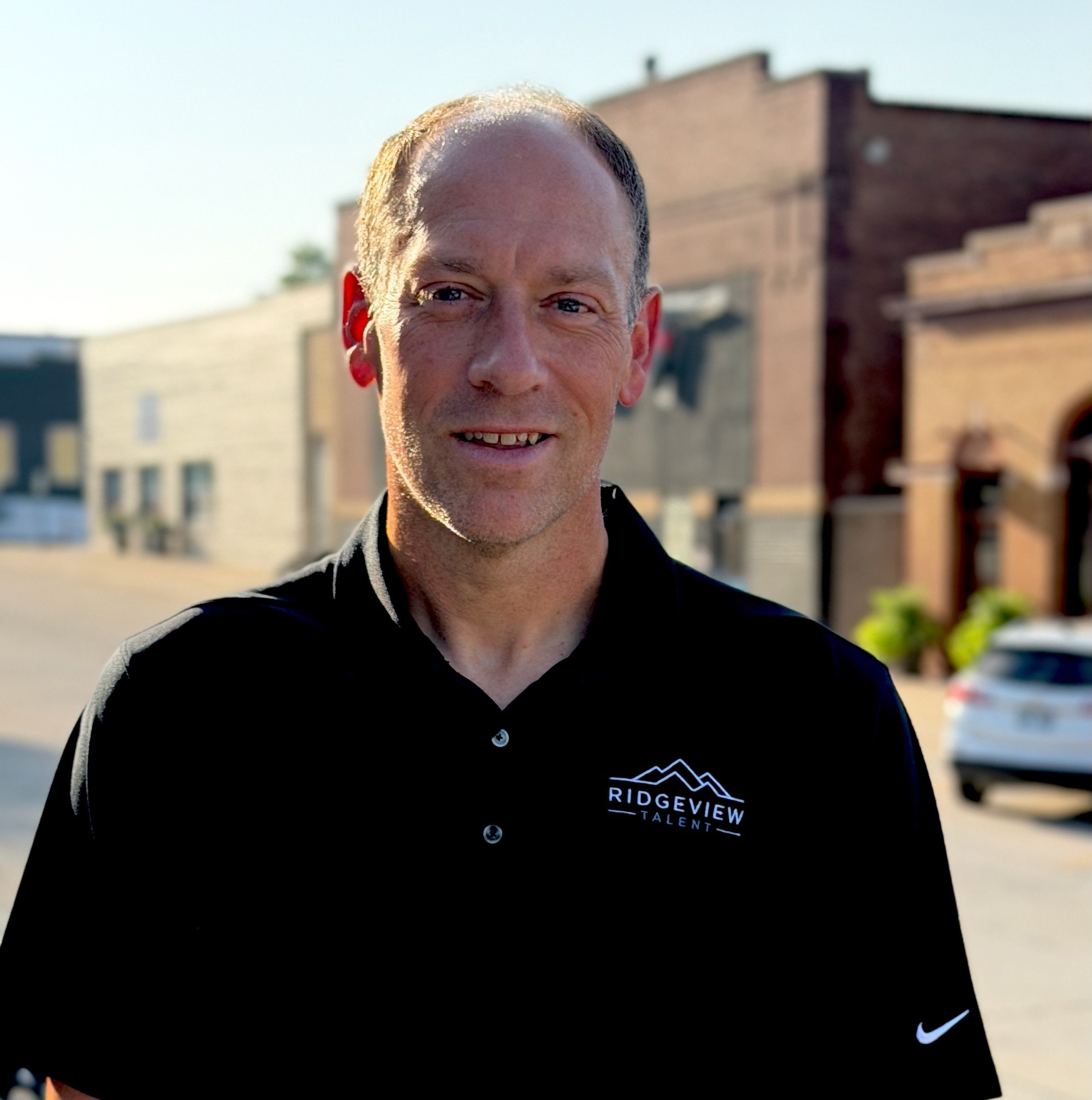 Close-up of smiling man with short hair wearing a black sports jacket with a Nike logo and a patch reading 'Ridgeview Talent,' standing outdoors with a building in the background.