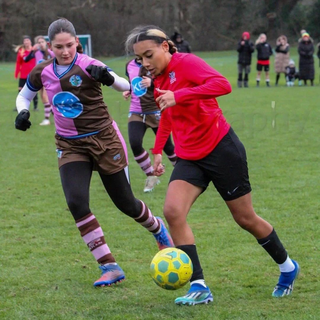 Two women playing soccer on a grassy field, with several people in the background watching