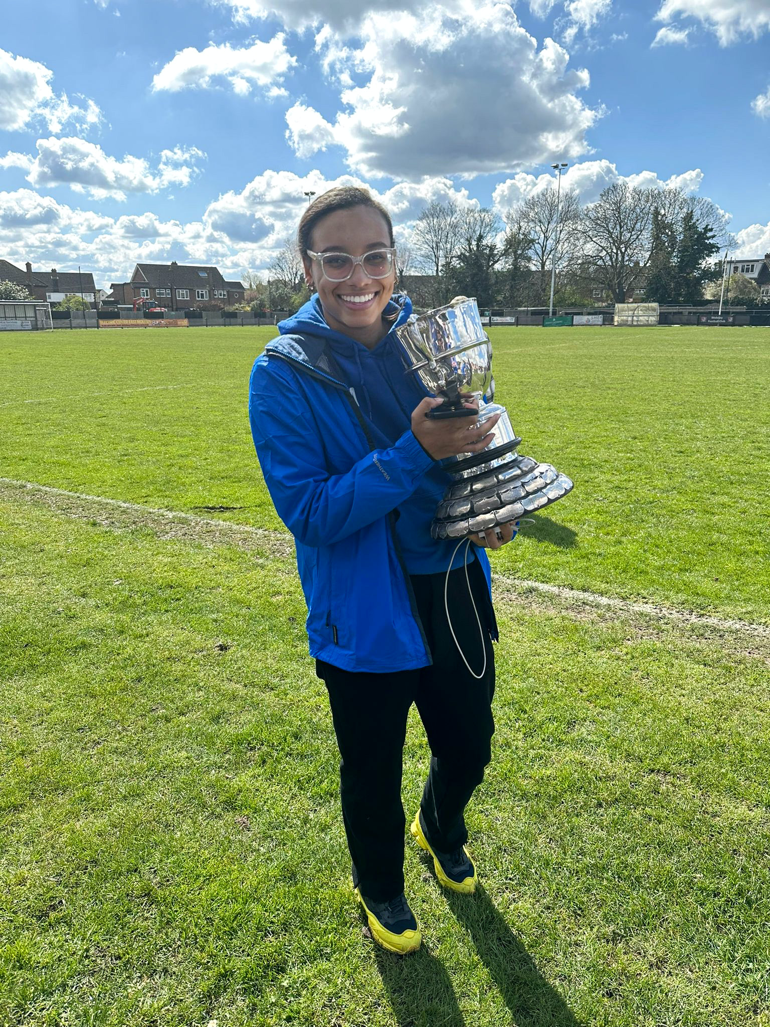 Socia founder Yasmine in a blue jacket holding a shiny silver trophy on a grassy field with houses and trees in the background under a partly cloudy sky.