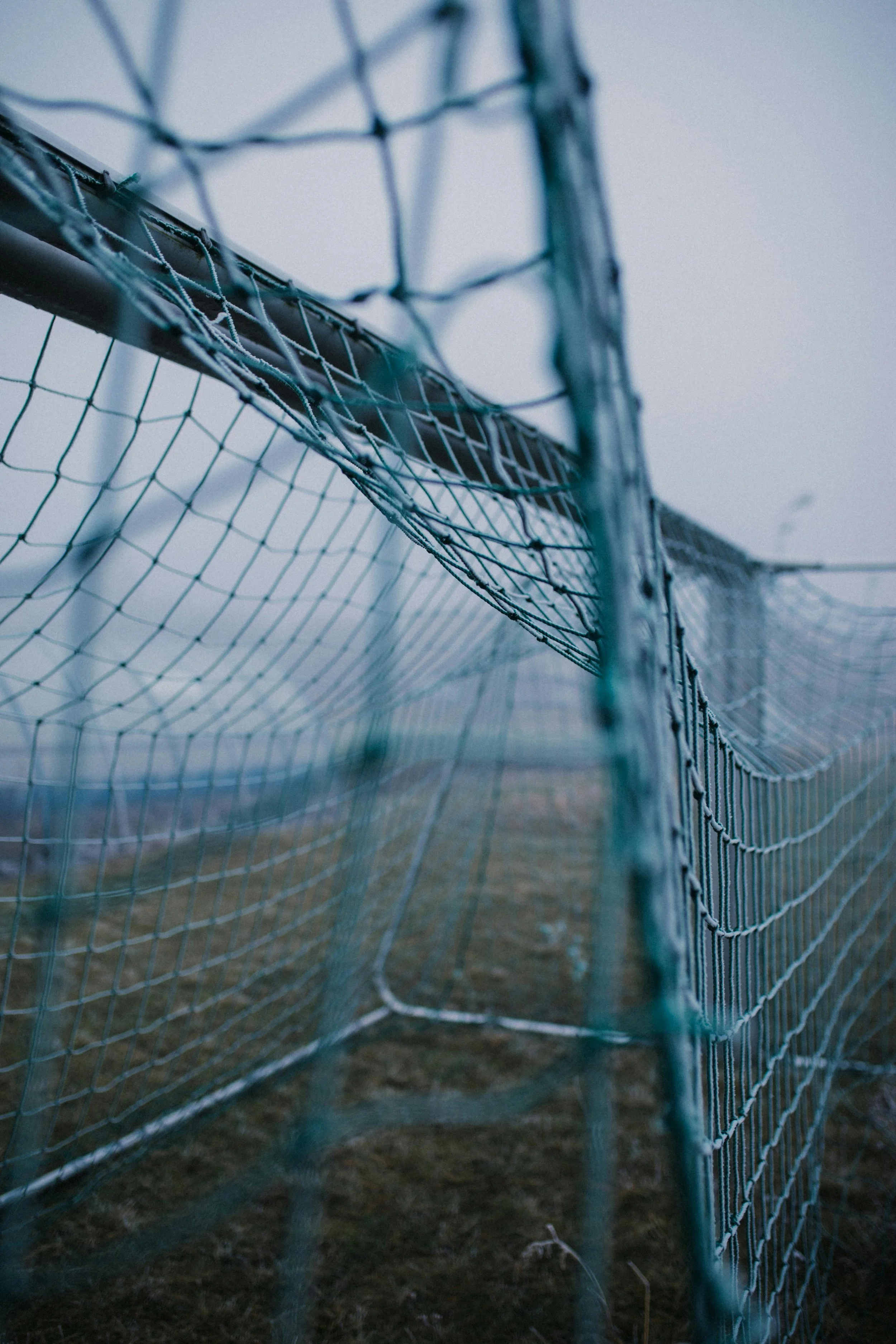 Close-up of a weathered soccer goal net attached to a metal post, with a foggy background and dull, muted colors.