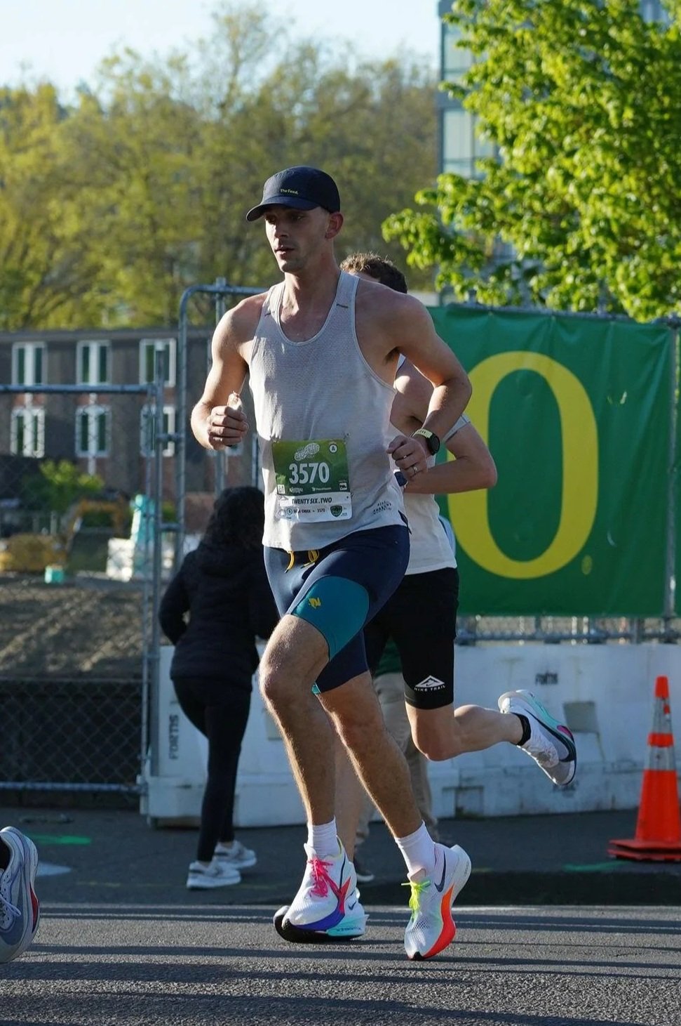 Male marathon runner wearing a black cap and white tank top, running outdoors during daytime with trees and a green Oregon Ducks banner in the background.