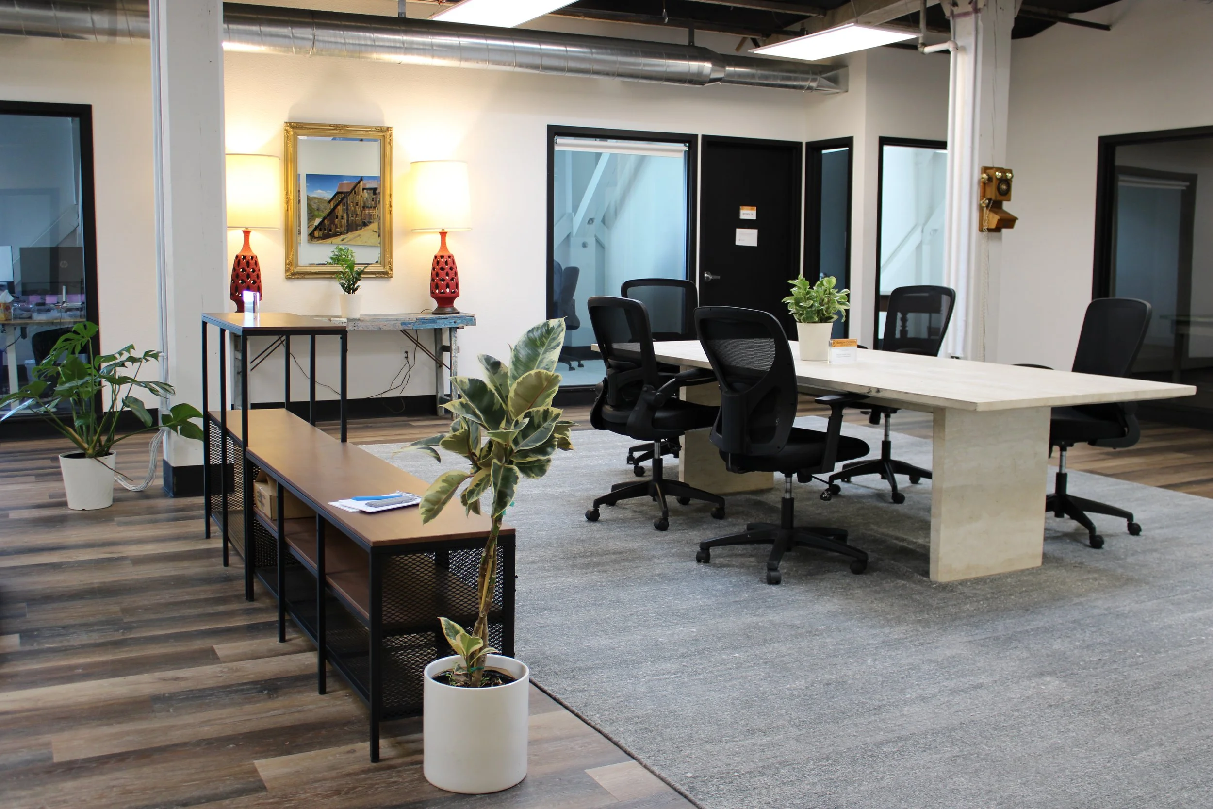Modern open-plan office with a long conference table, black office chairs, potted plants, artwork, and exposed ductwork