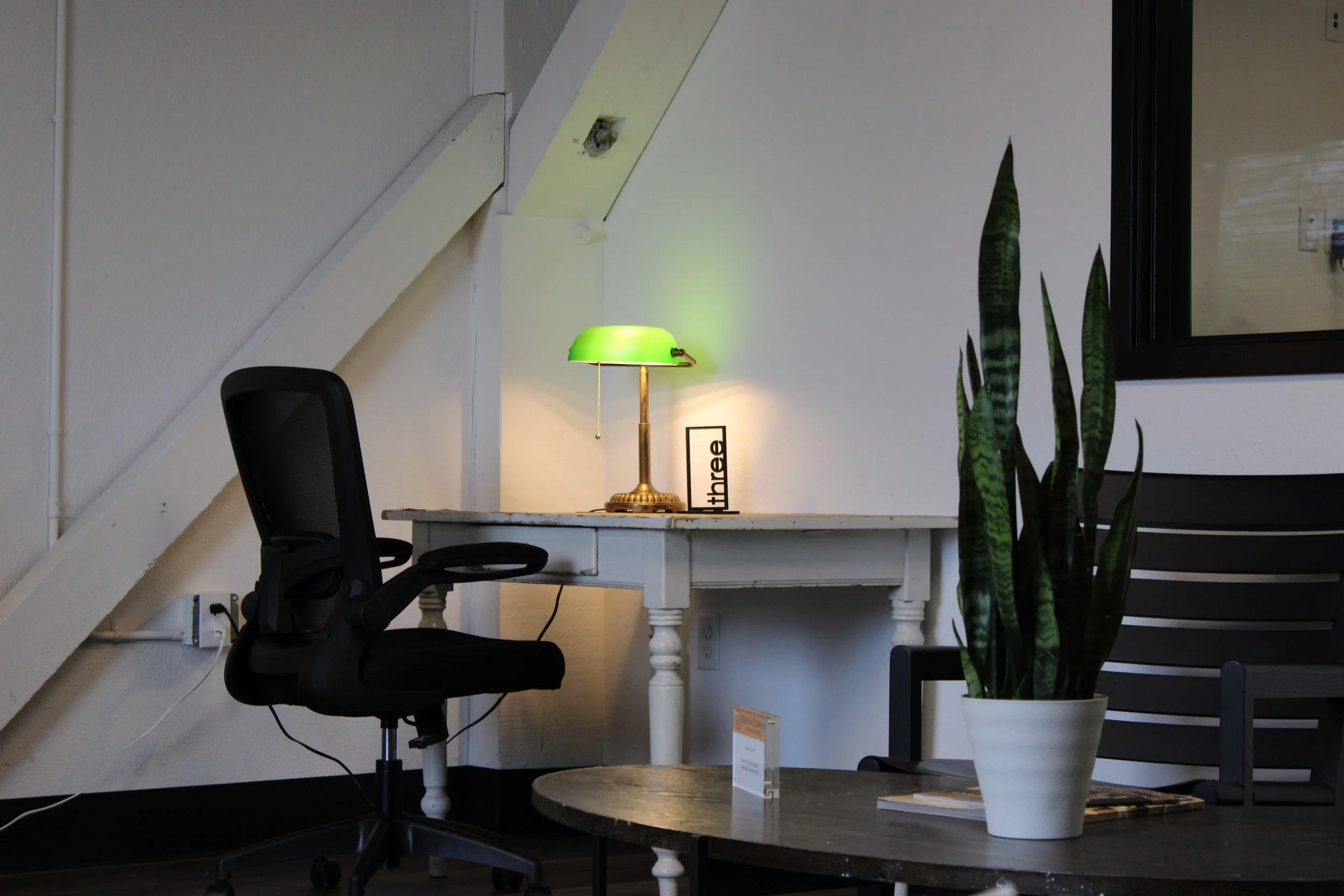 Modern home office with a black ergonomic chair, green banker’s lamp, white desk, snake plant, and coffee table