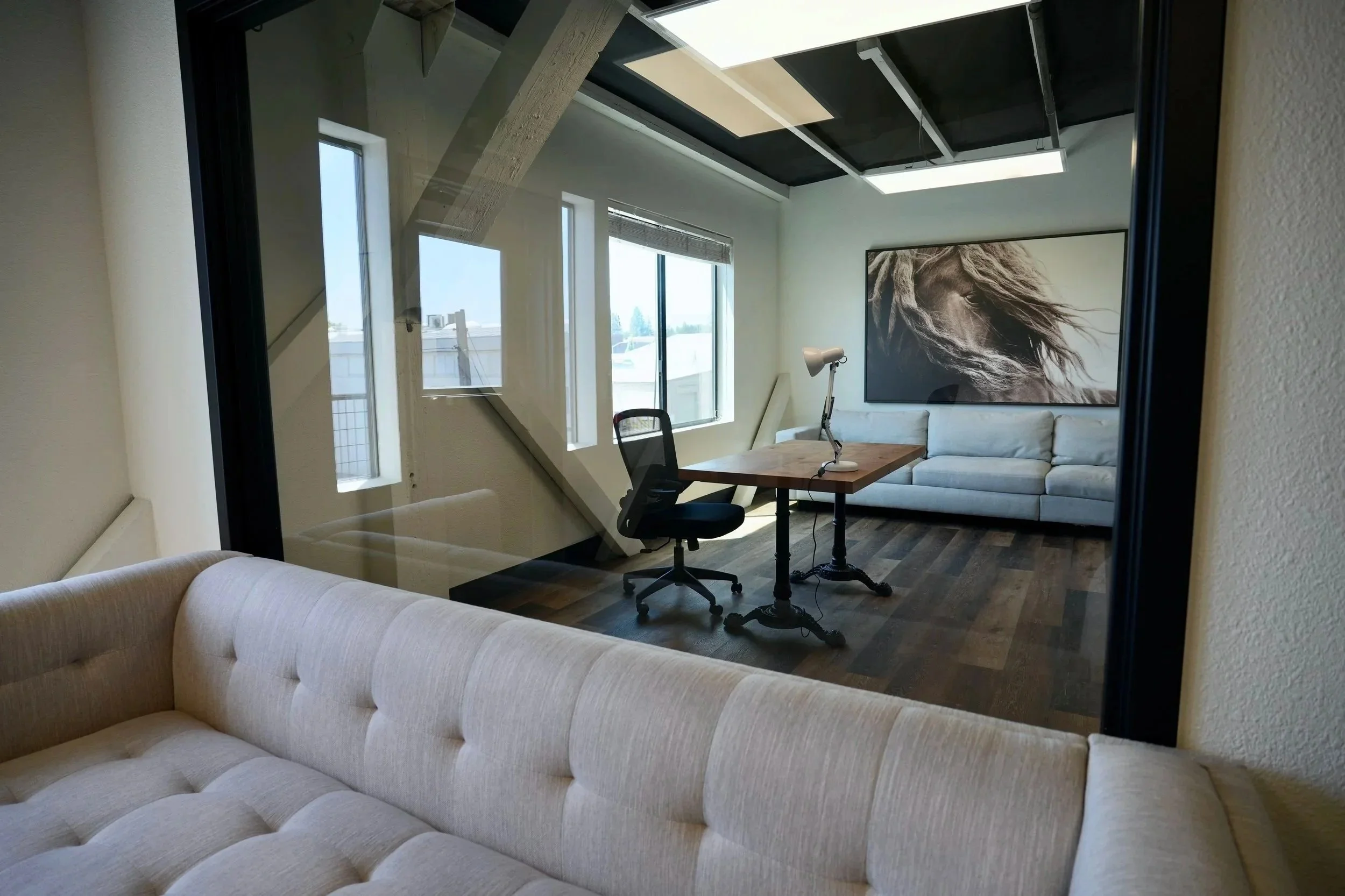 Bright modern office with a sectional sofa, wooden desk, office chair, floor lamp, large windows, and black-and-white portrait artwork
