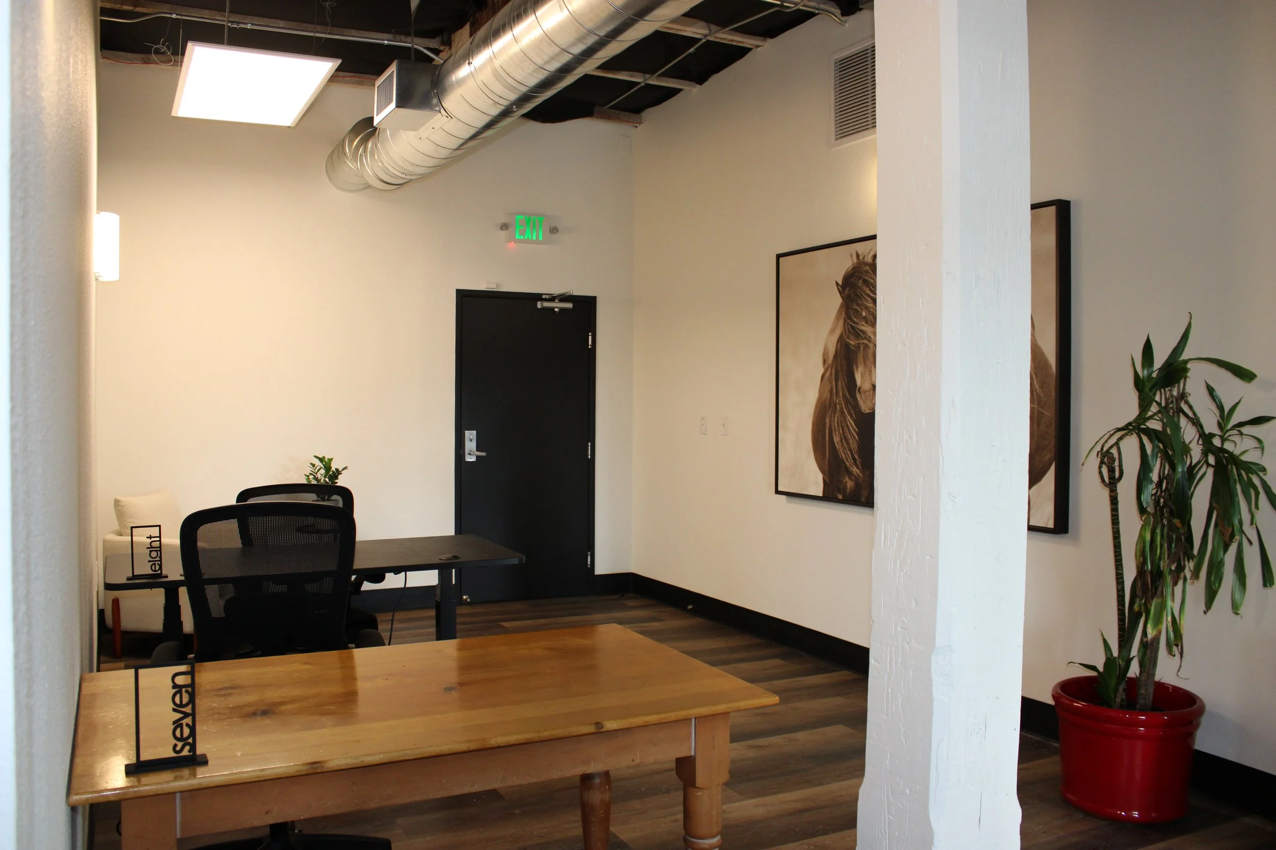 Modern office interior with wooden conference table, black office chairs, exposed ductwork, framed horse artwork, exit door, and potted plant