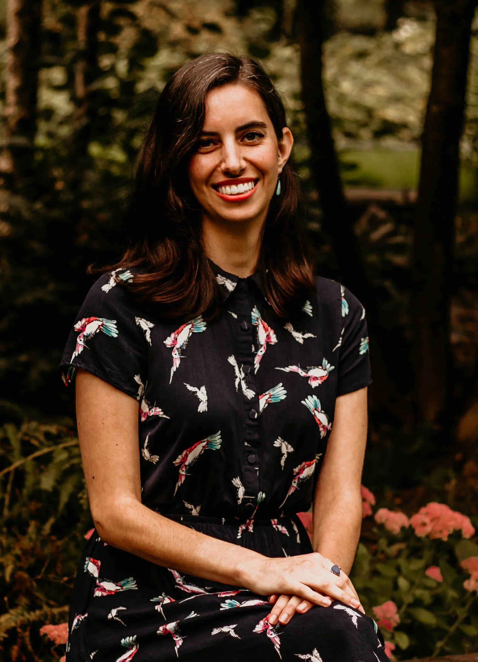 Photo of a woman with shoulder-length brown hair, tan skin, and a dark blue dress with hummingbirds. The woman is smiling, with a background of trees and flowers.