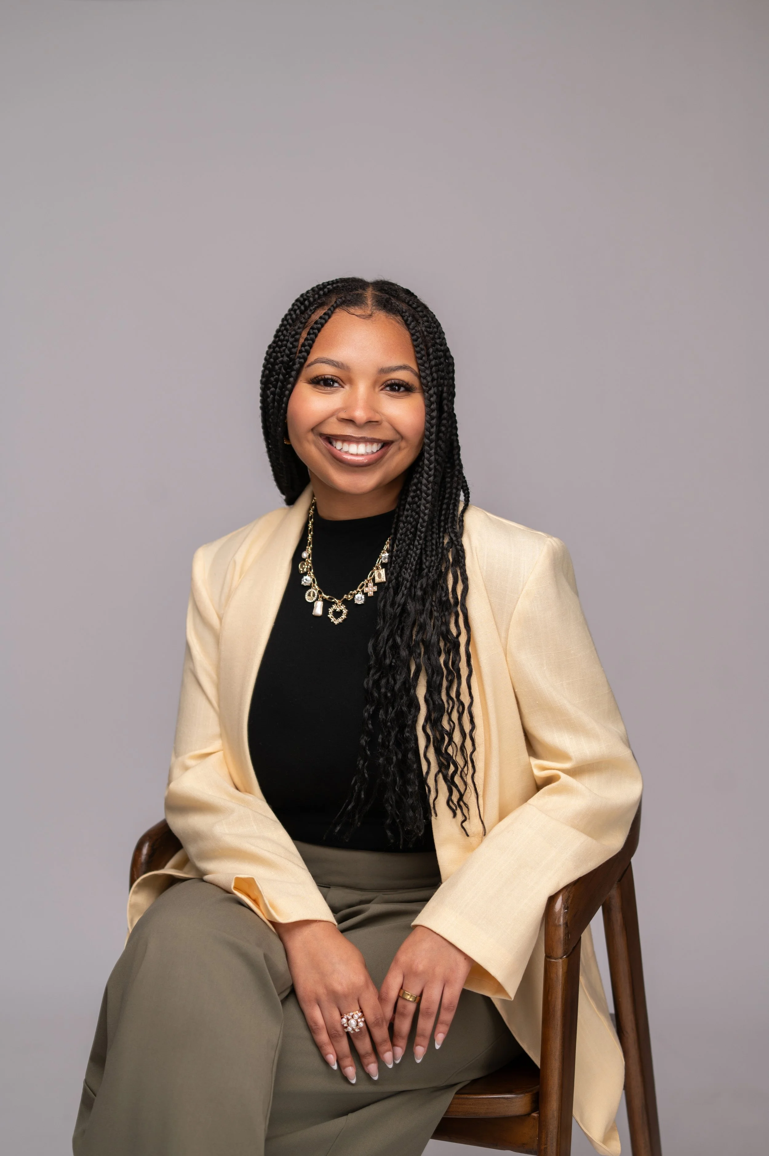 A woman sitting on a wooden chair against a neutral gray background, smiling at the camera. She has long braided hair, wears a black top, a beige blazer, and gold jewelry, including rings and necklaces.