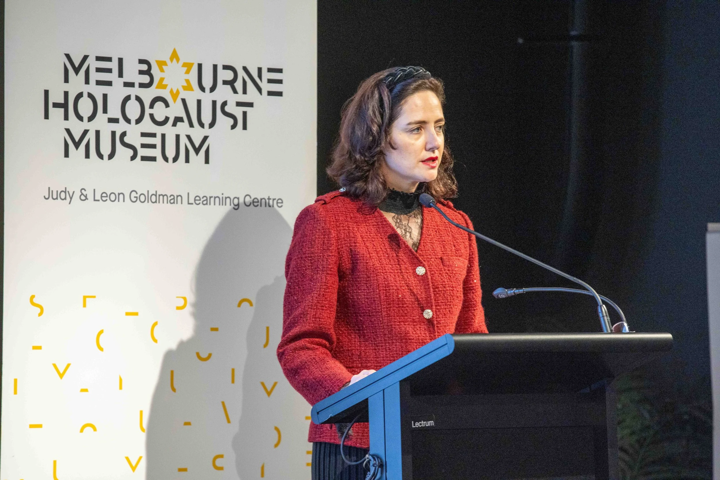 A woman in a red jacket and black dress speaking at a podium with a microphone, standing in front of a gray background with a white poster that reads 'Melbourne Holocaust Museum' and 'Judy & Leon Goldman Learning Centre'