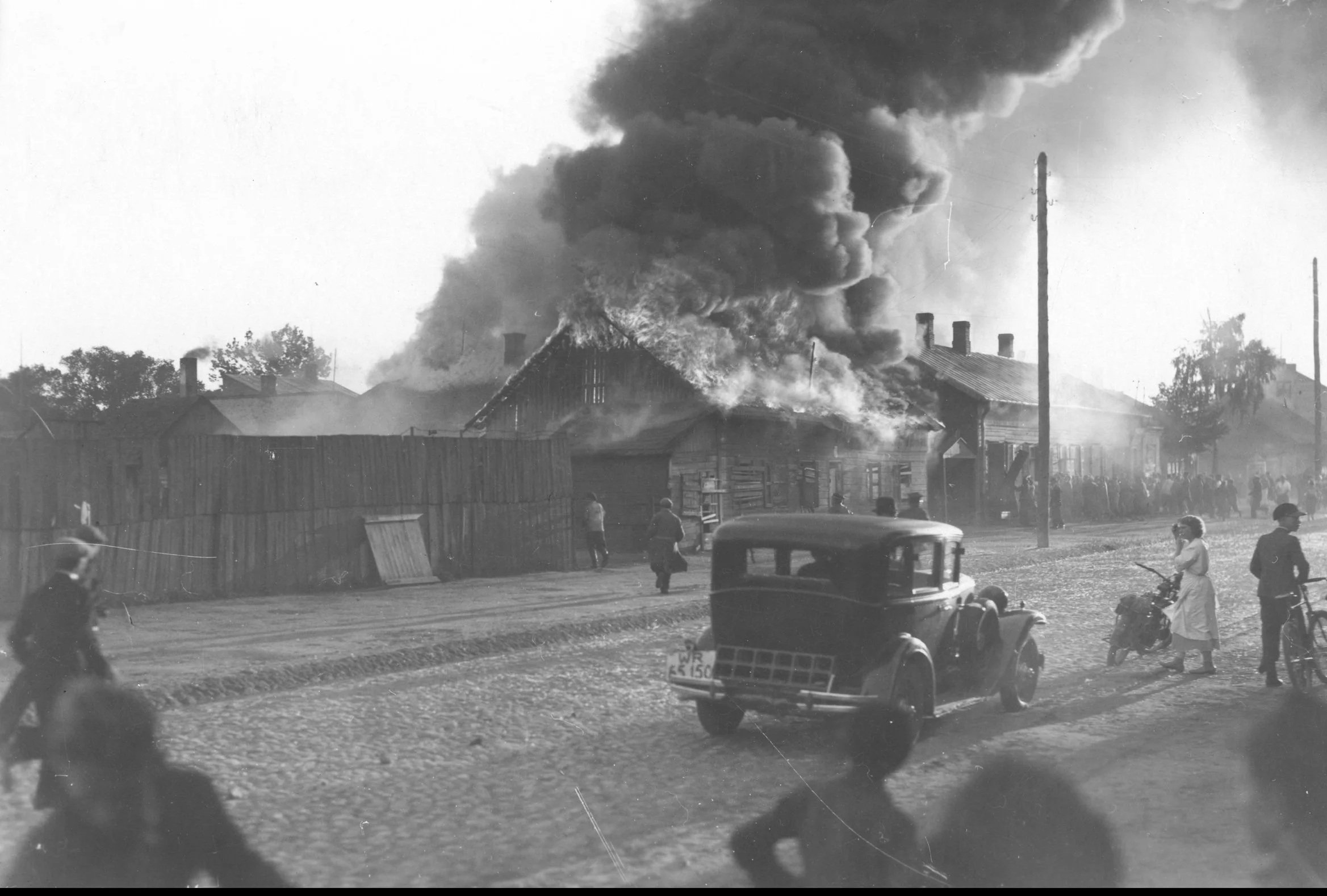 Black and white photograph of a house with a large fire and thick smoke billowing into the sky, with several children and vehicles in the foreground.