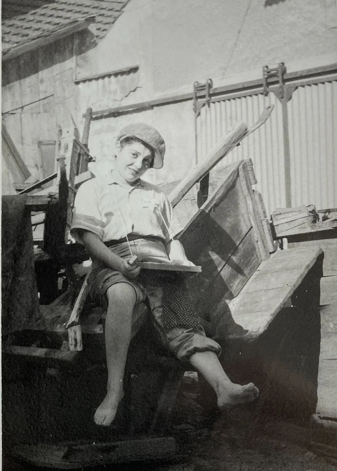 A young boy sitting on a wooden cart outdoors, smiling and holding a sailboat model, wearing a cap, short-sleeved shirt, shorts, and a scarf around his neck, with a background of a building with corrugated metal siding.