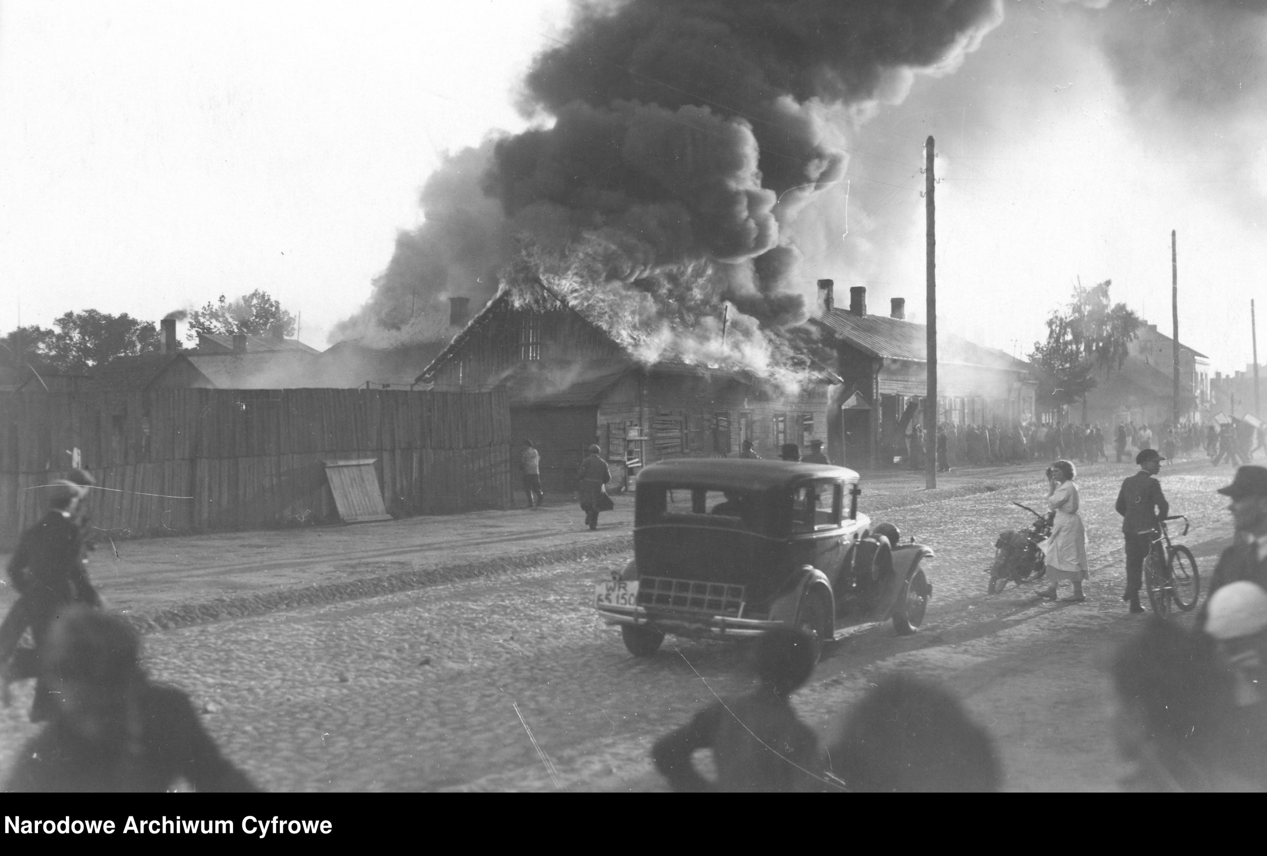 Black and white photo of a house on fire with thick smoke billowing from the roof, near utility poles and a street with children and cars in the foreground.
