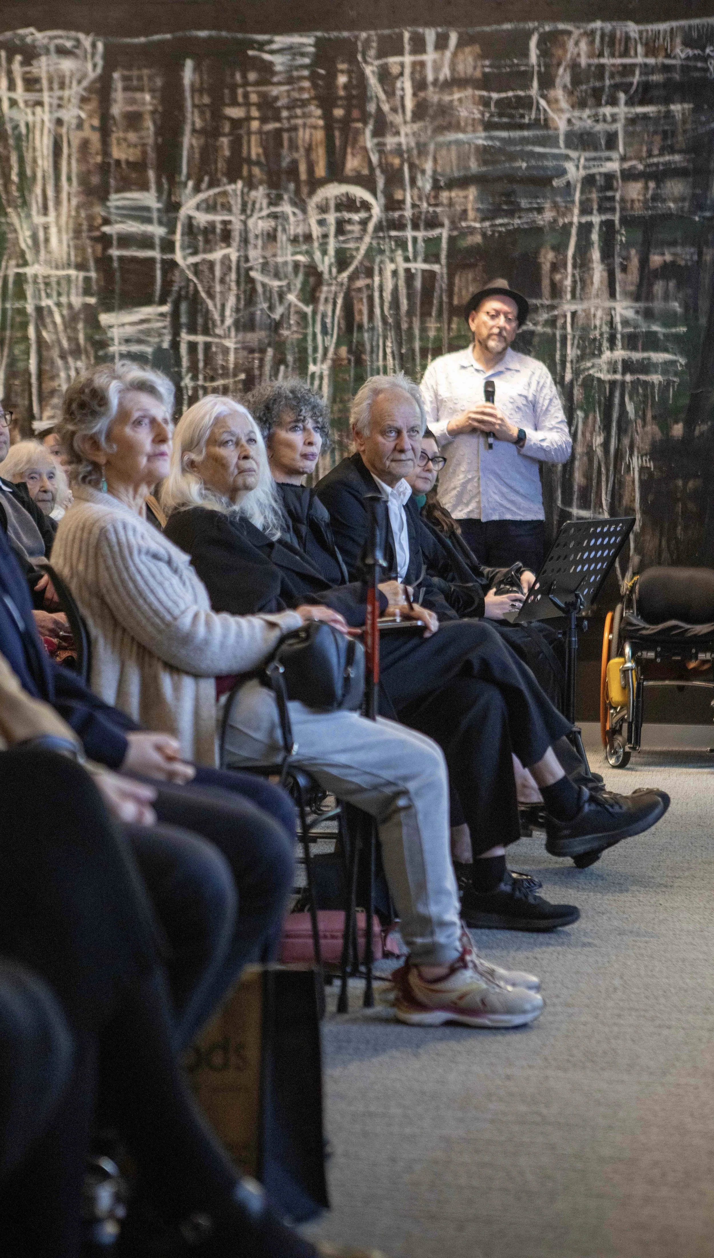 A group of elderly people seated indoors, listening to a speaker holding a microphone. The speaker is a man wearing a hat and a patterned shirt. The background features an abstract mural on the wall.