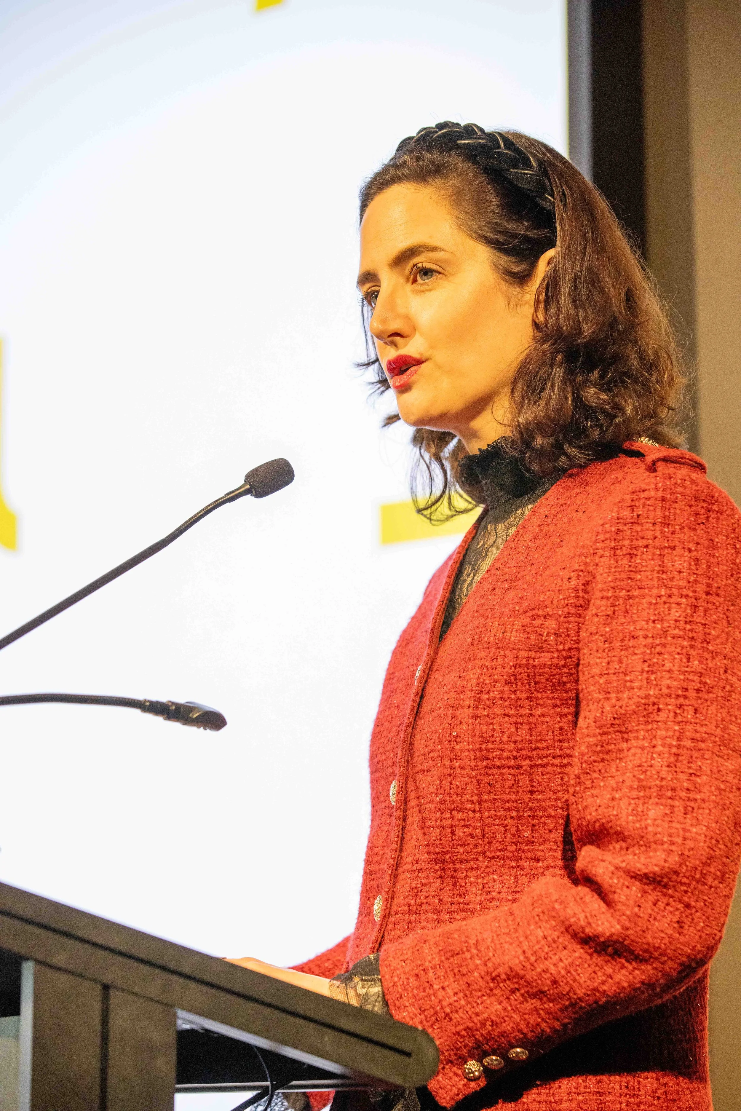 A woman with wavy brown hair and red lipstick giving a presentation at a podium, wearing a red textured blazer with gold buttons and a black top underneath.