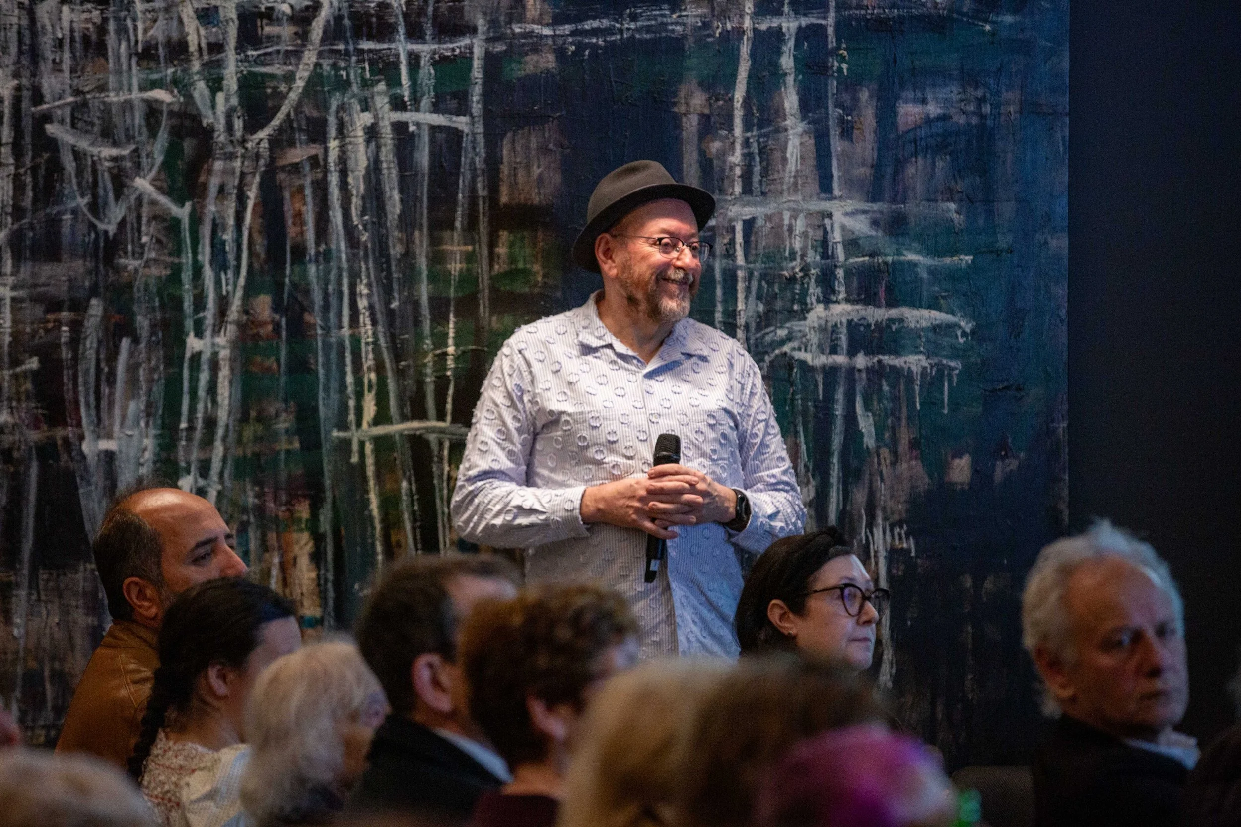 A man wearing glasses, a dark hat, and a printed shirt is smiling and holding a microphone while standing in front of a colorful abstract painting. He is surrounded by seated audience members at an indoor event.