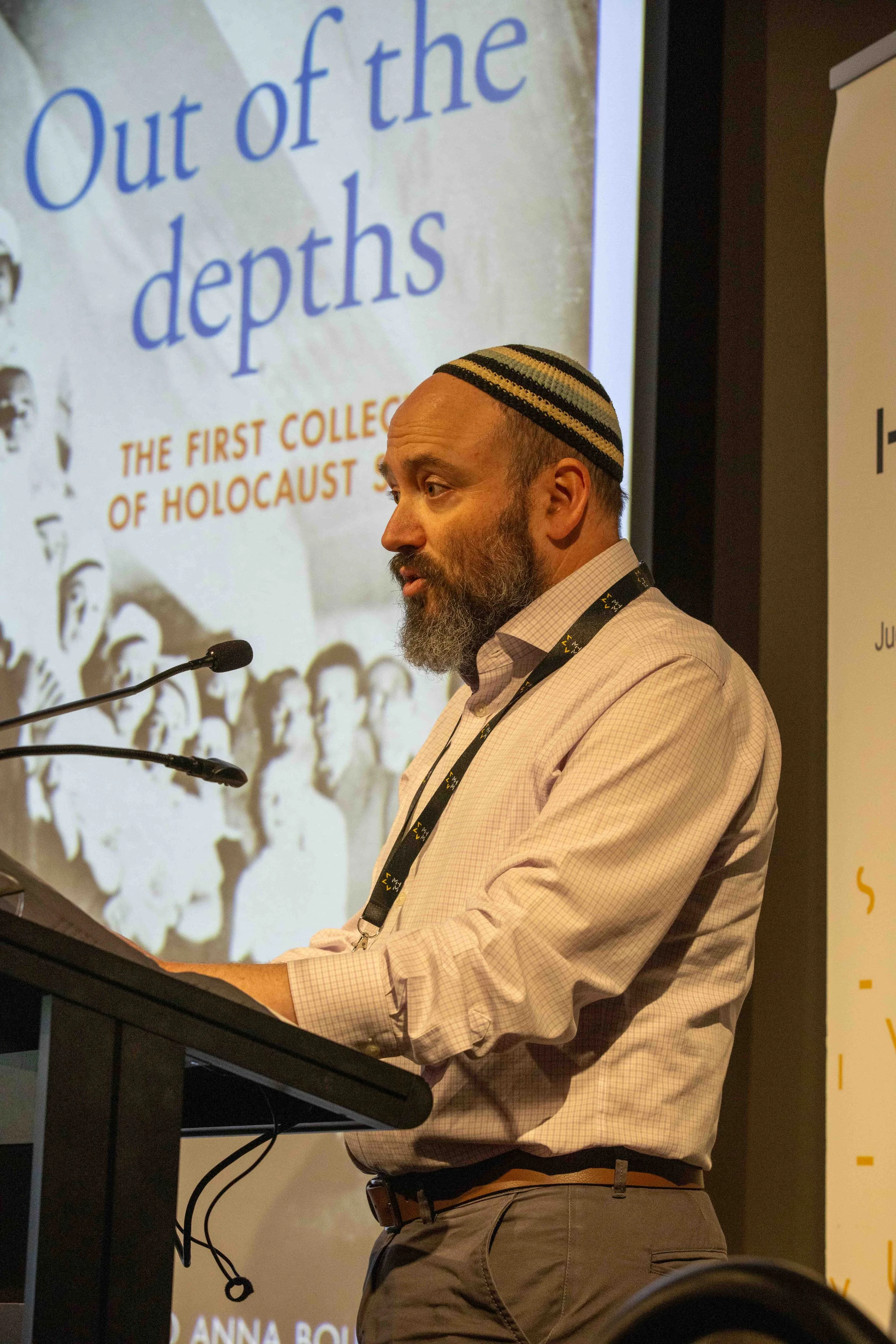 A man wearing a kippah and a light plaid shirt is standing at a podium with a microphone. Behind him, there is a screen displaying a presentation slide with the title 'Out of the depths' and the subtitle 'The first collection of Holocaust stories.'