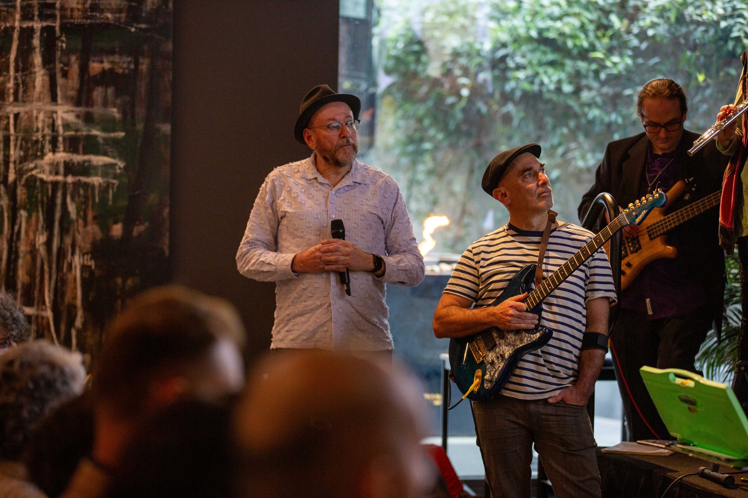 Group of musicians performing indoors, with a man in striped shirt holding a guitar, another man with glasses and a hat, and a woman playing a bass guitar, surrounded by trees and greenery outside.