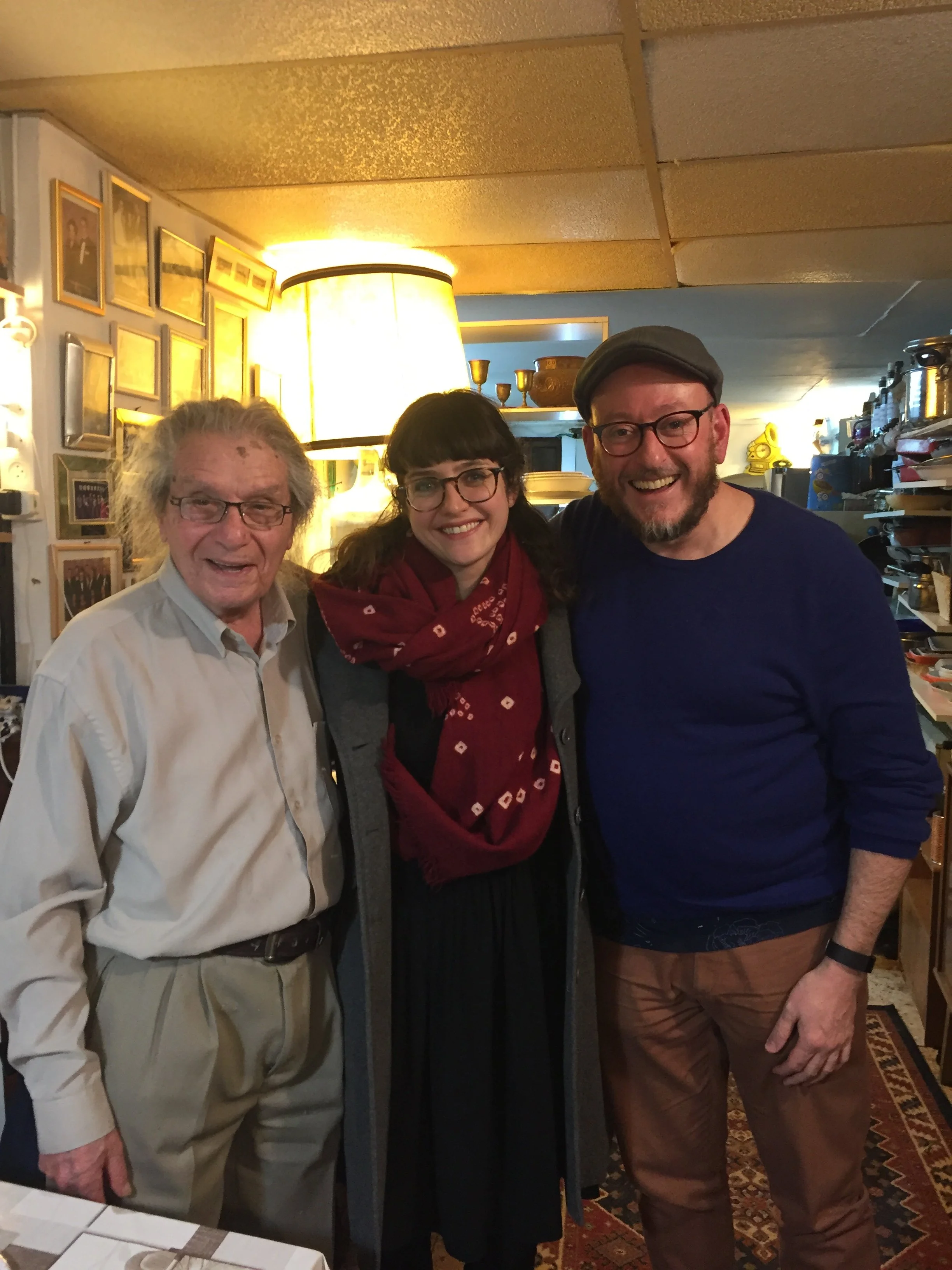 Three people smiling for a photo in a warmly lit room with framed pictures on the wall and various decorative items on shelves.
