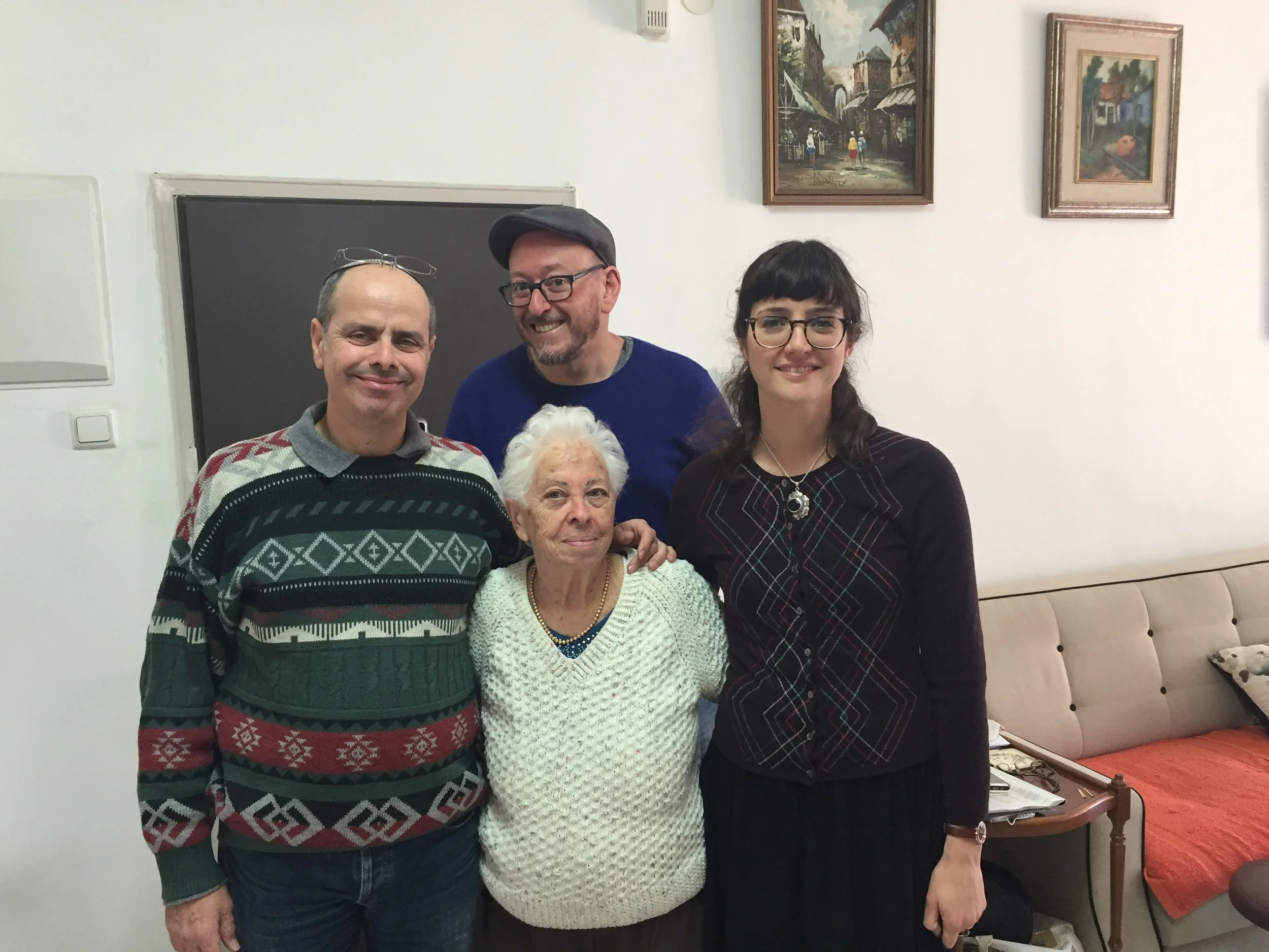 Four people standing together inside a room, smiling at the camera. The room has a white wall with framed paintings and a television. The group includes an older woman with white hair, two middle-aged women, and a middle-aged man wearing glasses and a hat.