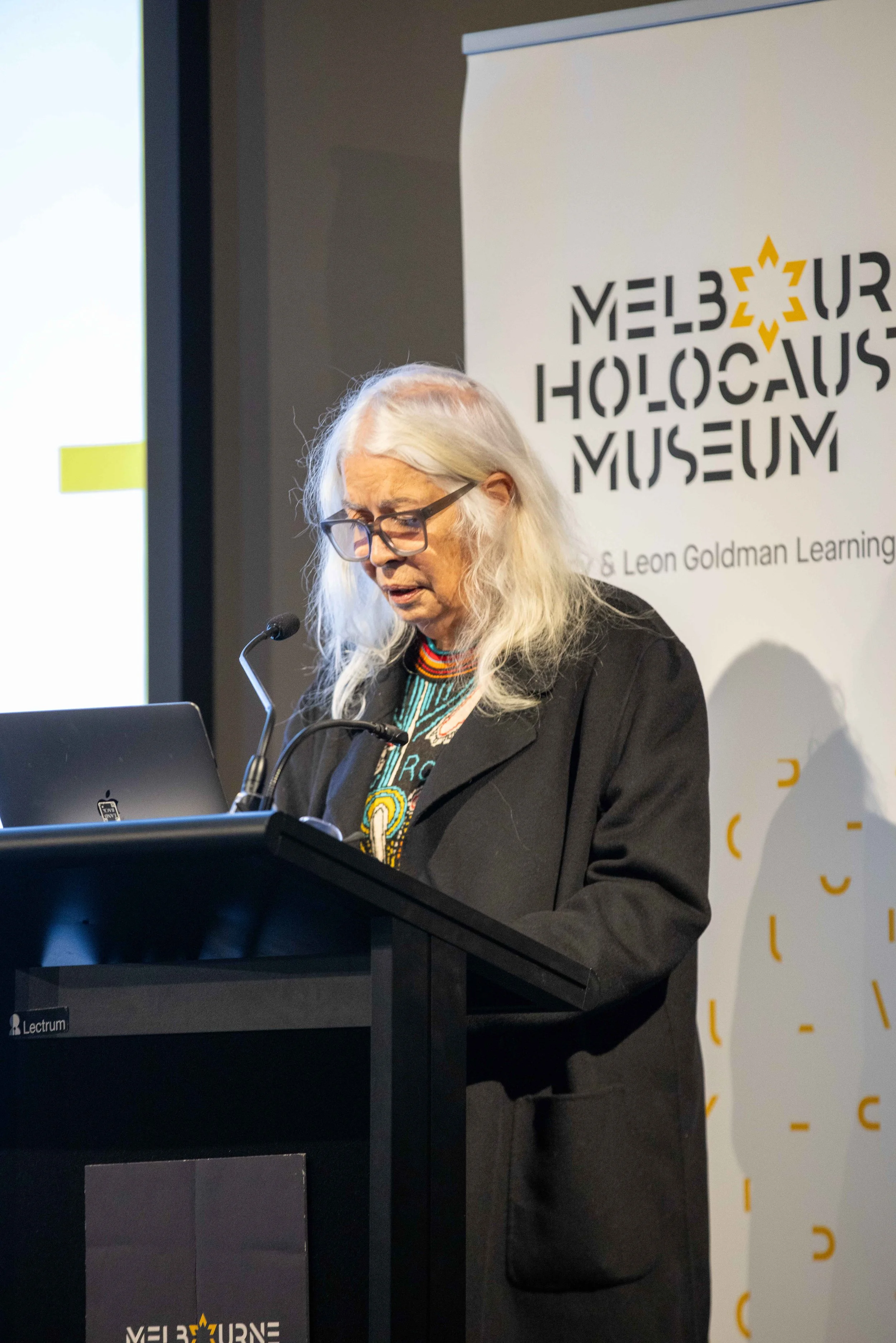 An elderly woman with long white hair and glasses, wearing a black jacket over a colorful patterned shirt, is speaking at a podium during a presentation or event at the Melbourne Holocaust Museum, with a logo and text on a banner behind her.