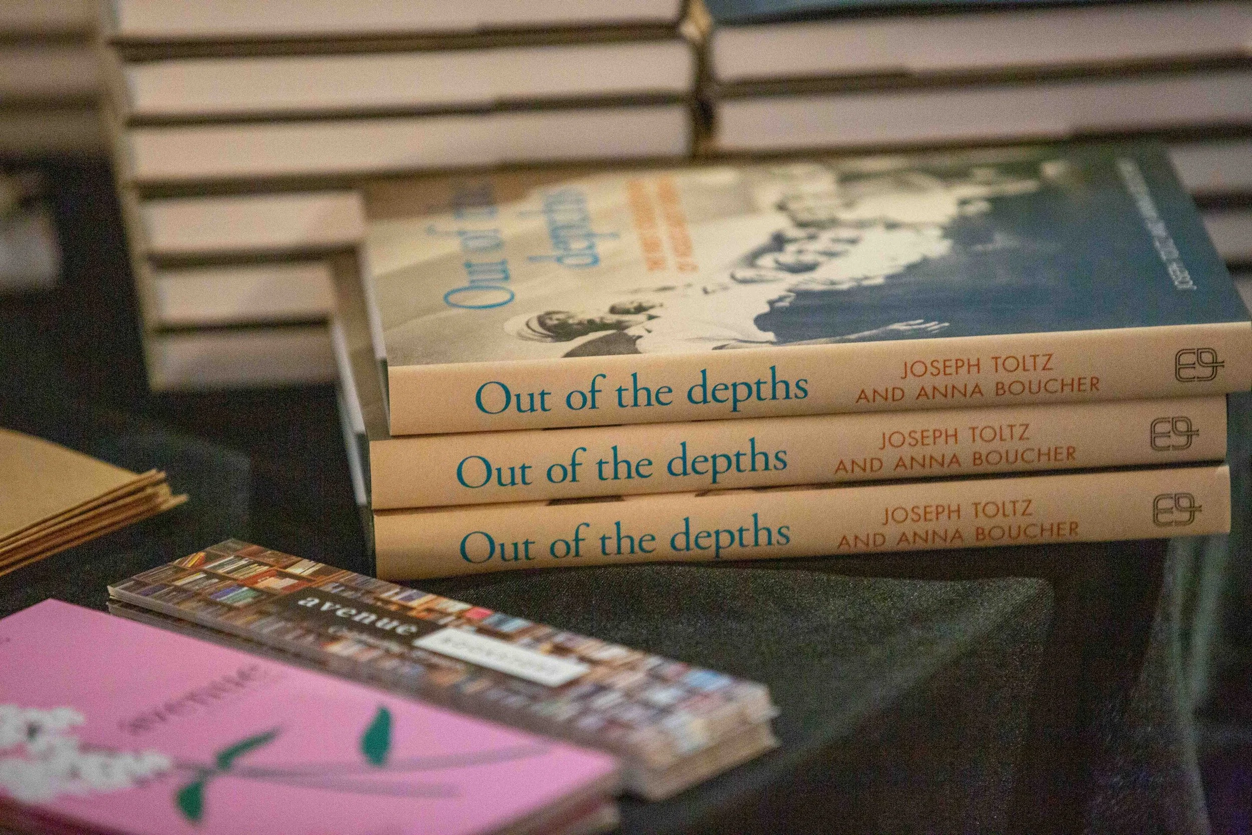 Stack of books titled 'Out of the depths' by Joseph Toltz and Anna Boucher on a black surface with other books and papers nearby.