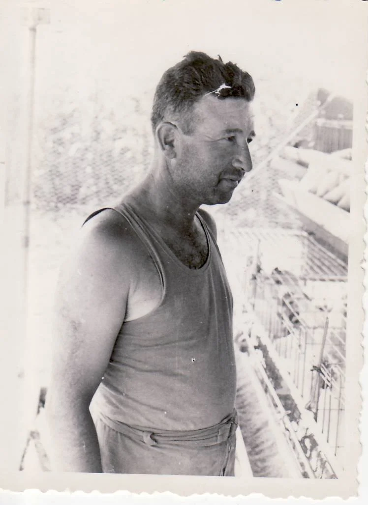 A black and white photograph of a man in a sleeveless shirt standing on a balcony or outdoor area, looking to his right. In the background, there are stacks of logs or firewood.
