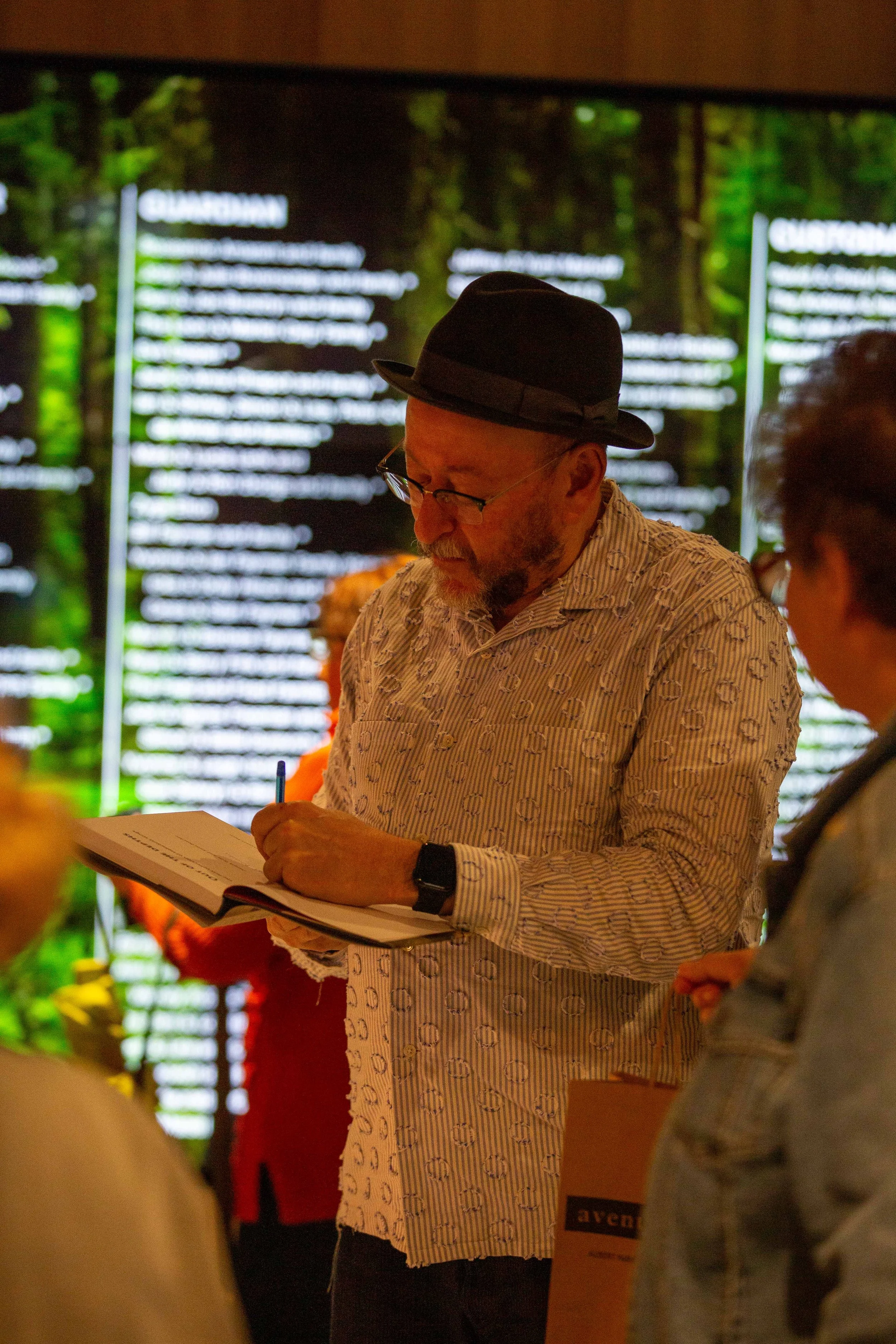 A man wearing glasses, a black hat, and a patterned shirt is reviewing a document in a crowded indoor setting with a large digital display in the background showing a list of names.