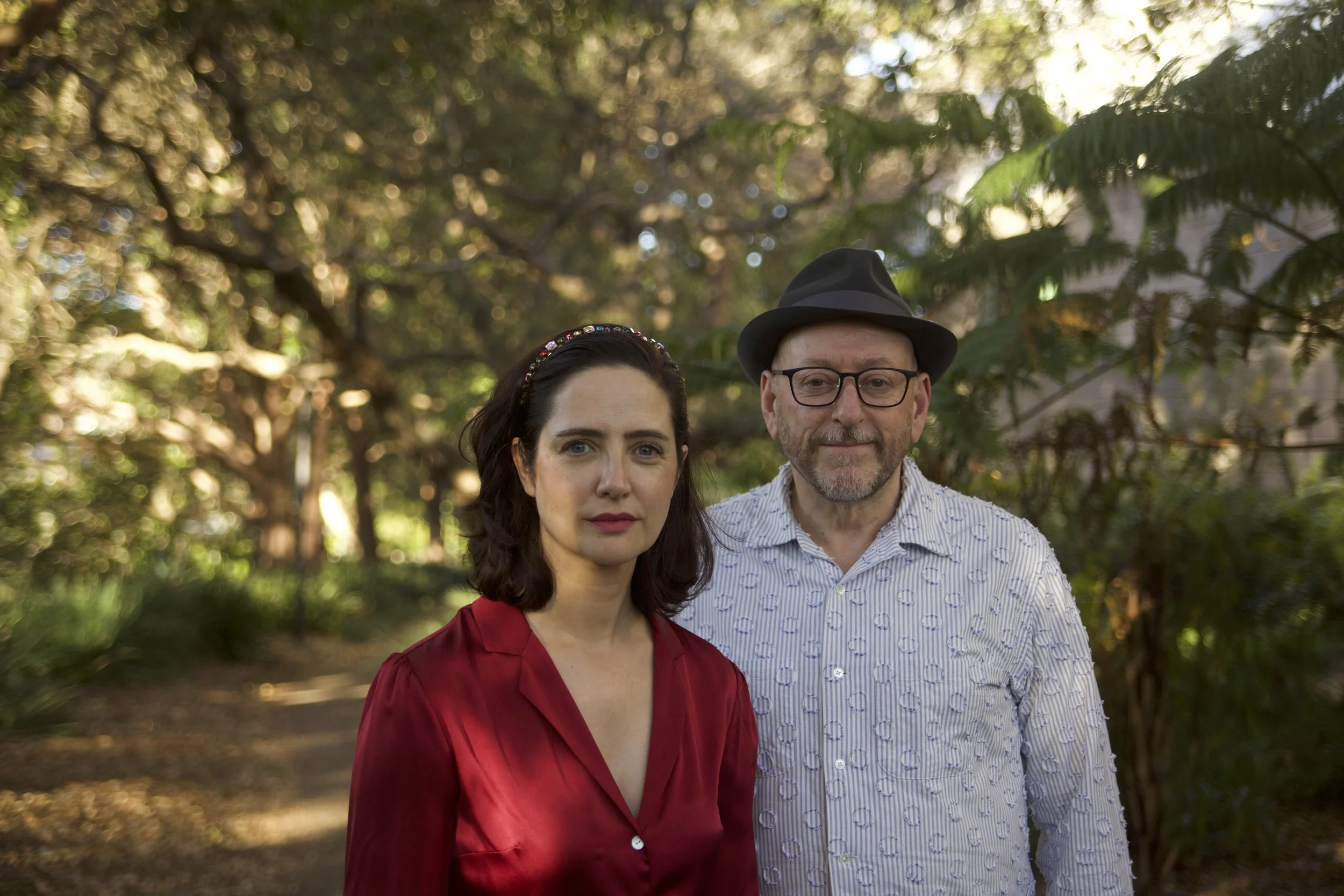 A woman with dark hair and a man with glasses and a hat standing outdoors among trees during daytime.