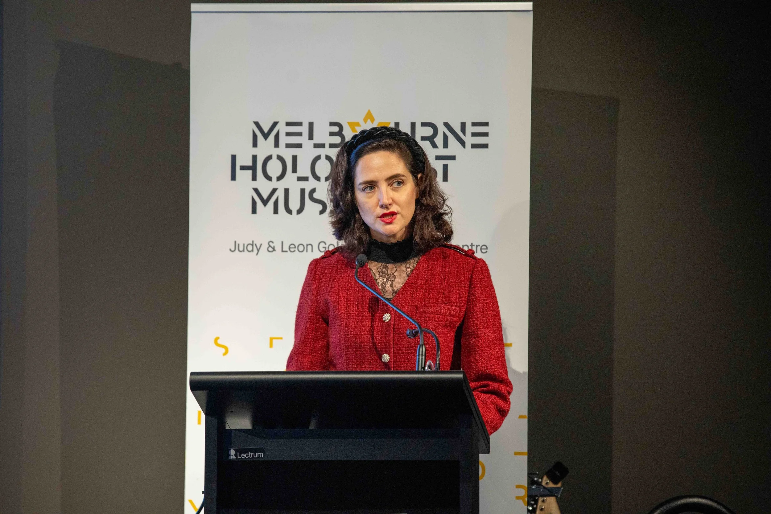 A woman in a red jacket and black lace top standing at a lectern, speaking at an event with a banner in the background that reads 'Melbourne Holoc...'.