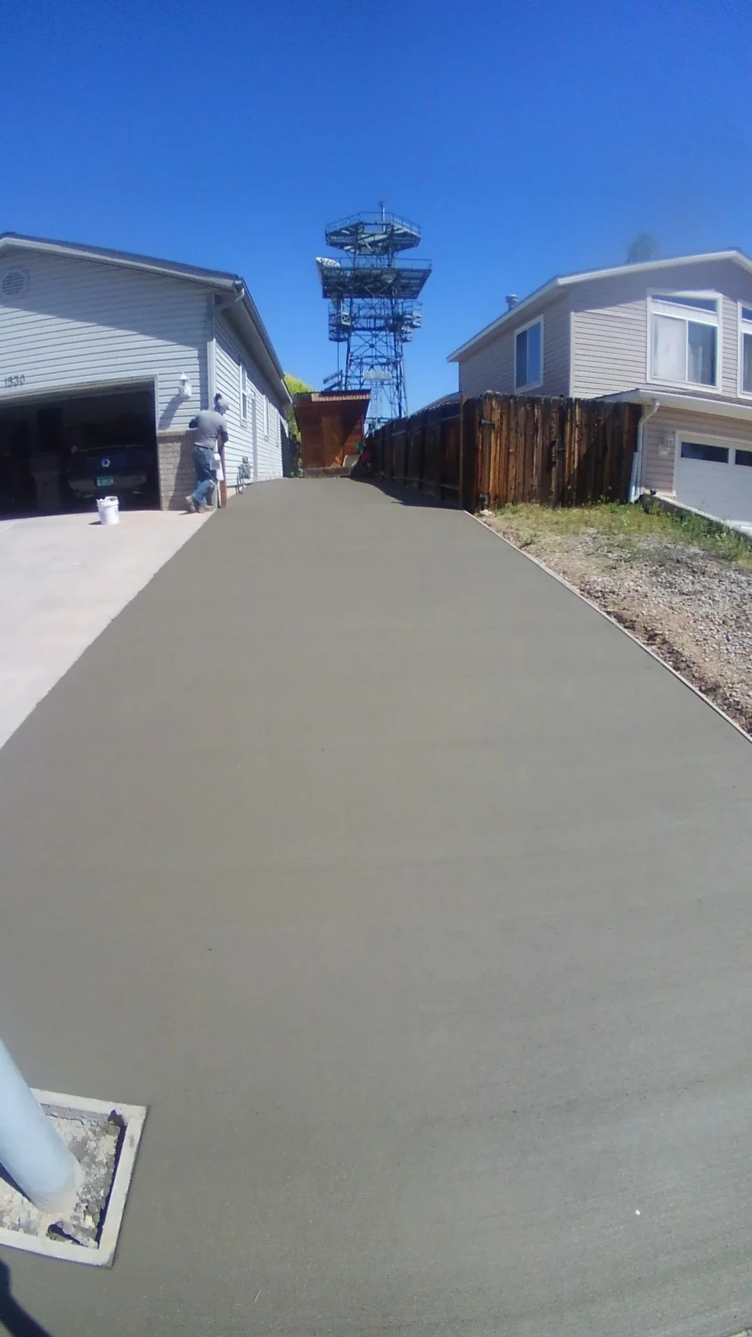 Freshly poured concrete driveway leading to a house with a man on a ladder, and a wooden fence under a clear blue sky.