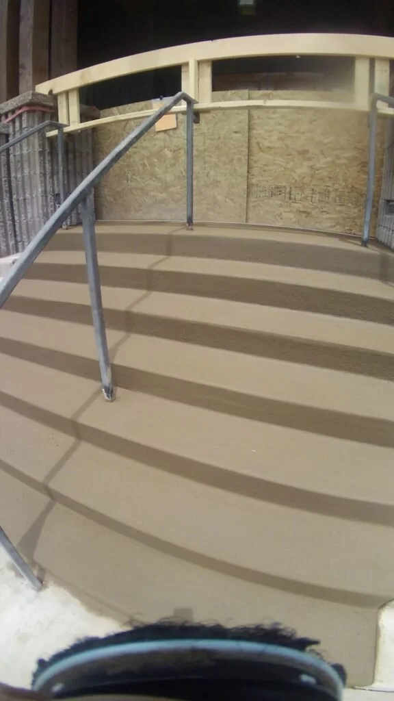 Empty outdoor ramp with metal railings and a curved wooden overhead barrier, casting shadows on the beige surface.