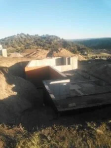A construction site with partially built concrete structures in a hilly area with trees and clear blue sky.