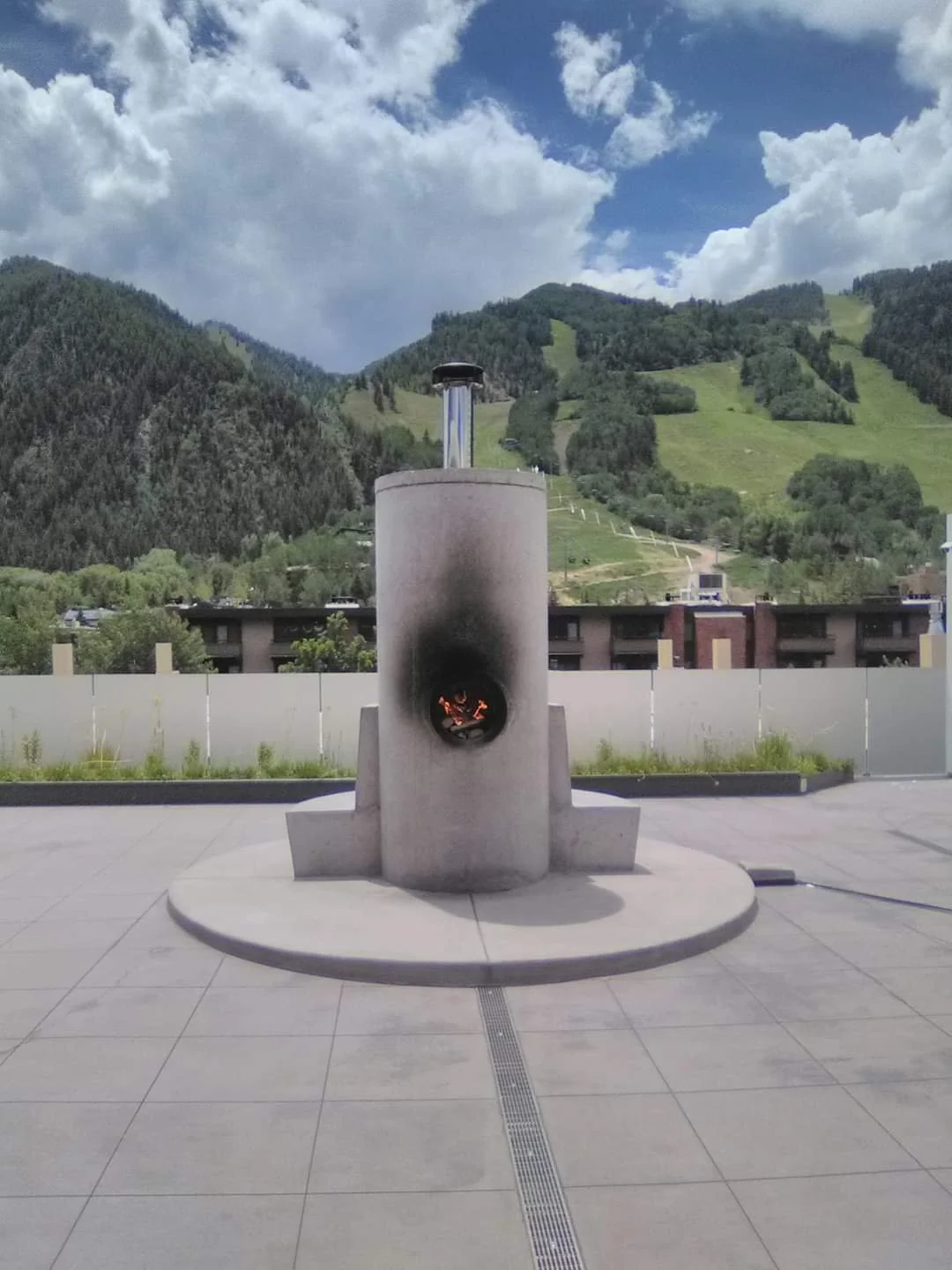 A modern outdoor pizza oven with a chimney, set on a concrete patio, with scenic green hills and a partly cloudy sky in the background.