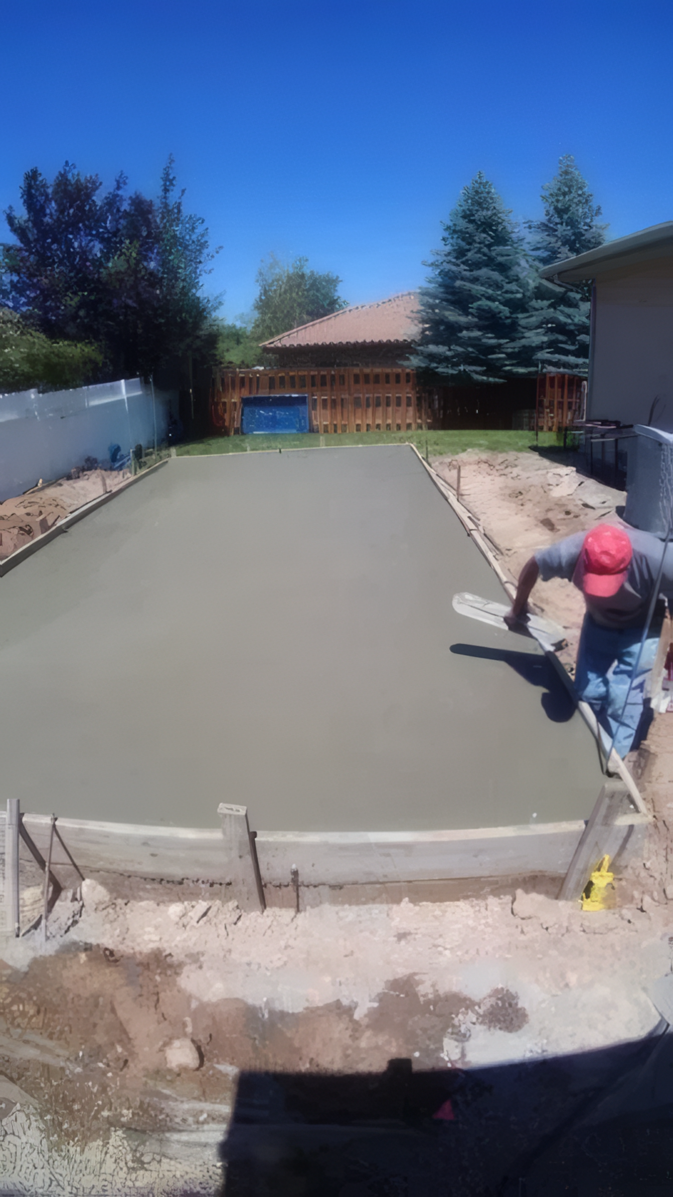 A person wearing a pink hat and gray shirt is smoothing wet concrete in a backyard patio area, with a wooden fence, trees, and a house in the background.