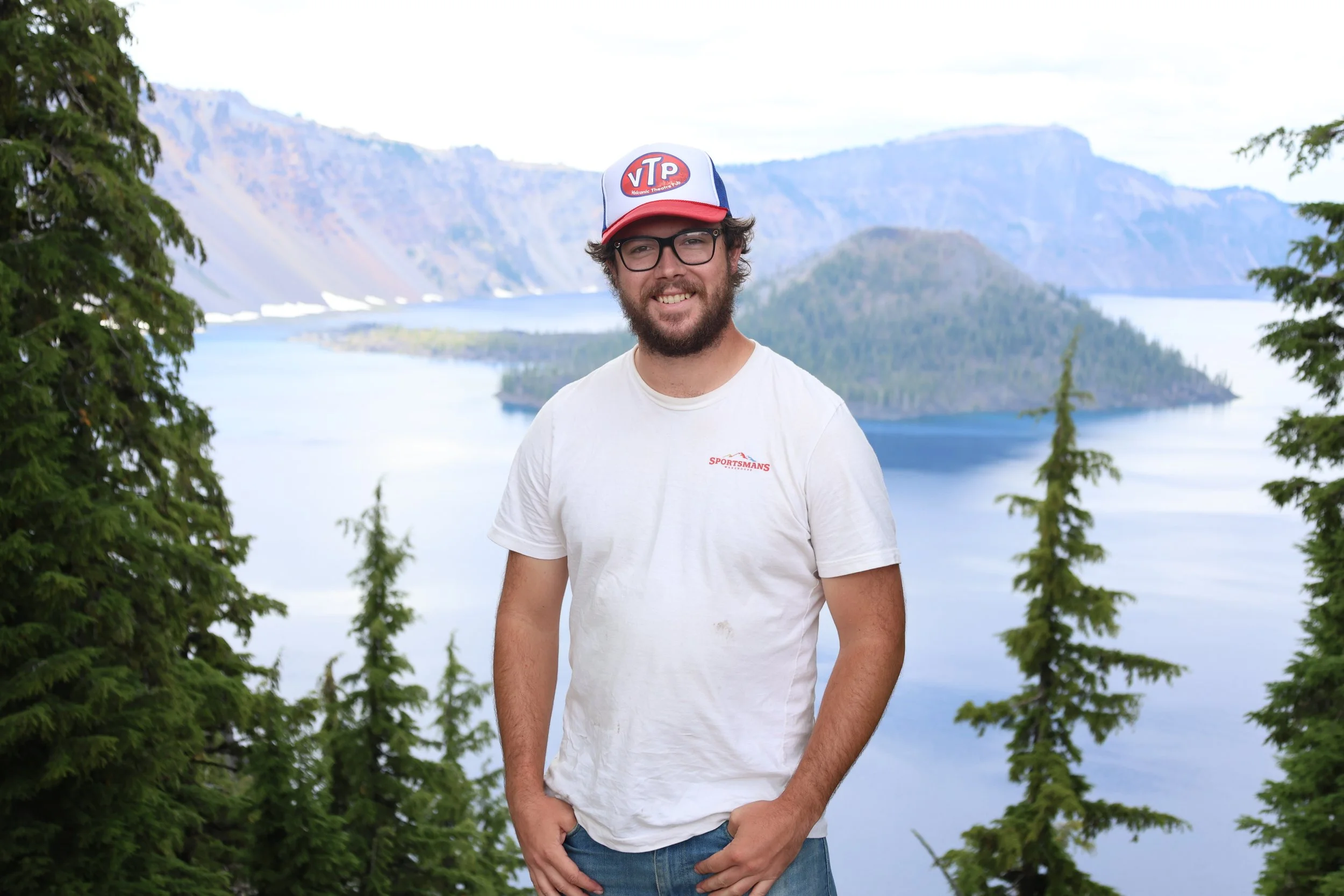 A young man with glasses, a beard, and curly hair, wearing a white t-shirt and a baseball cap, standing outdoors near a lake with trees and mountains in the background.