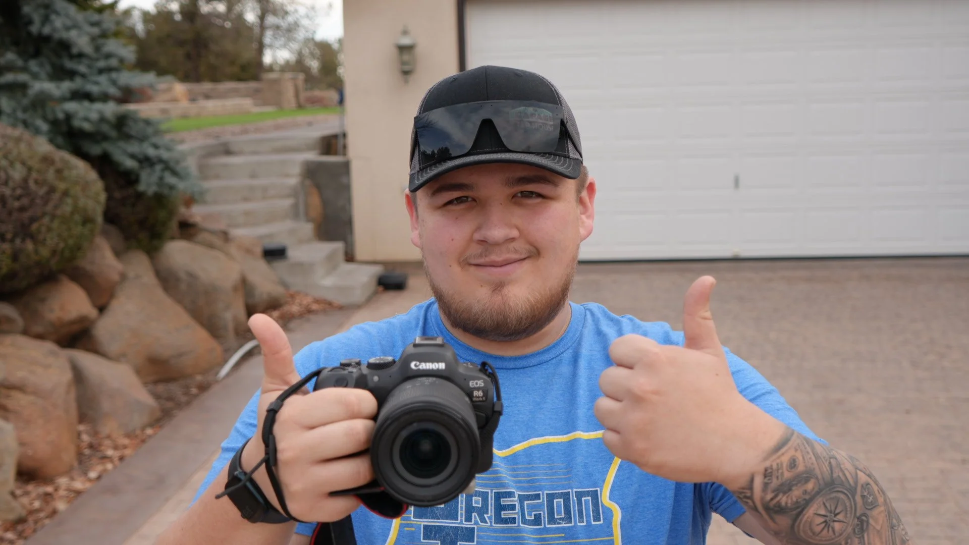 Young man in blue T-shirt holding a Canon camera and smiling with thumbs up outside near a garage.