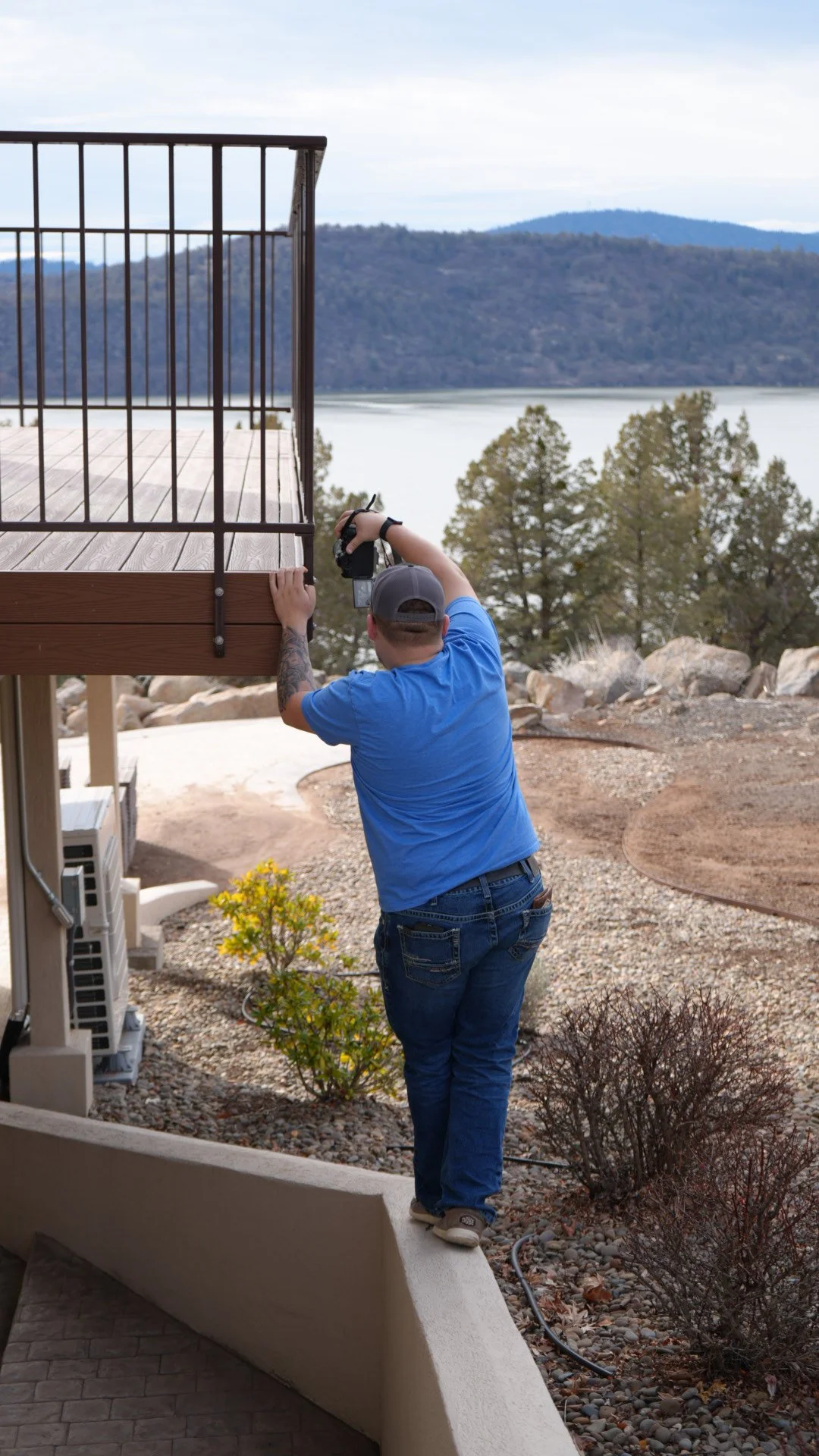A man in a blue shirt and baseball cap is taking a photograph from a balcony overlooking a lake and mountains in the distance.
