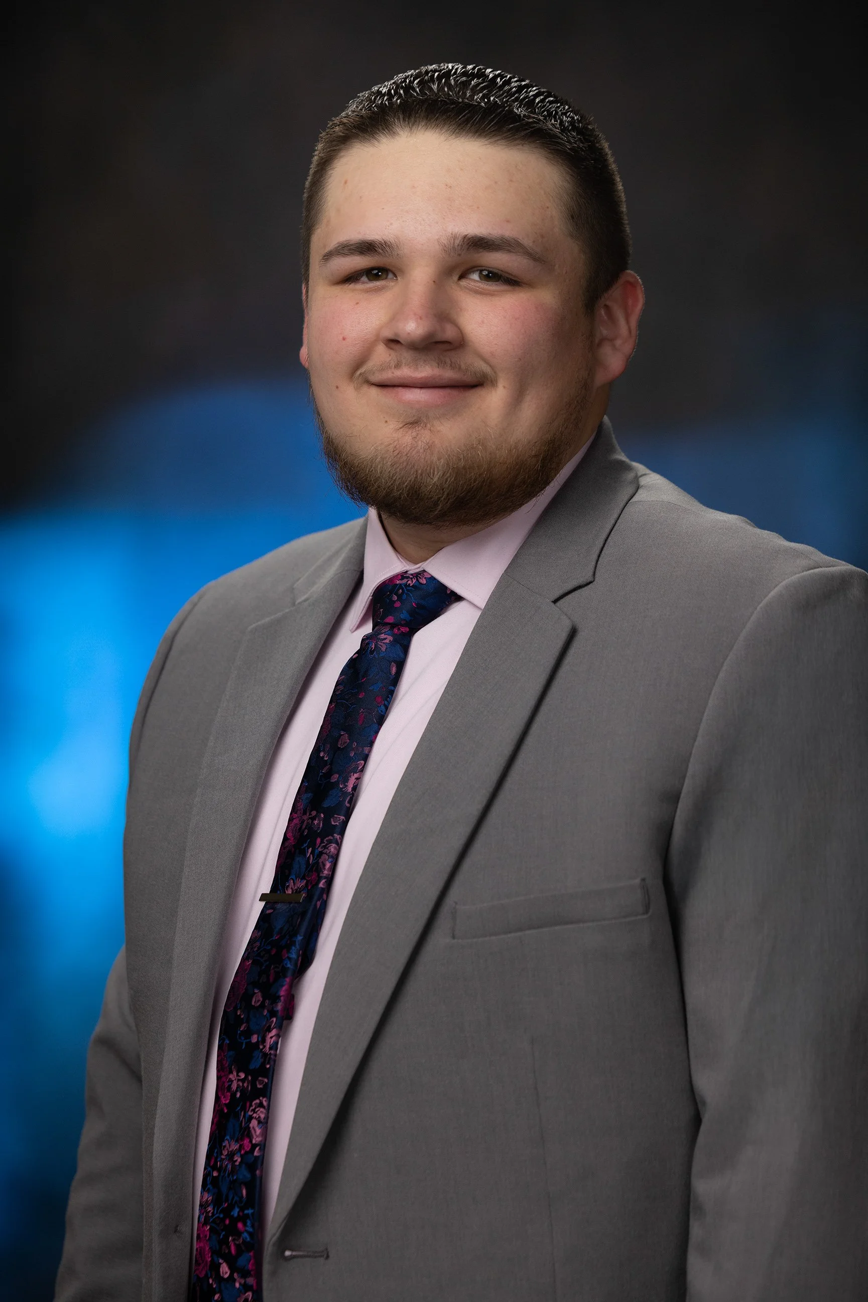A young man with a beard and short hair dressed in a gray suit, pink shirt, and floral tie, posing for a professional portrait against a dark background with blue lighting accents.
