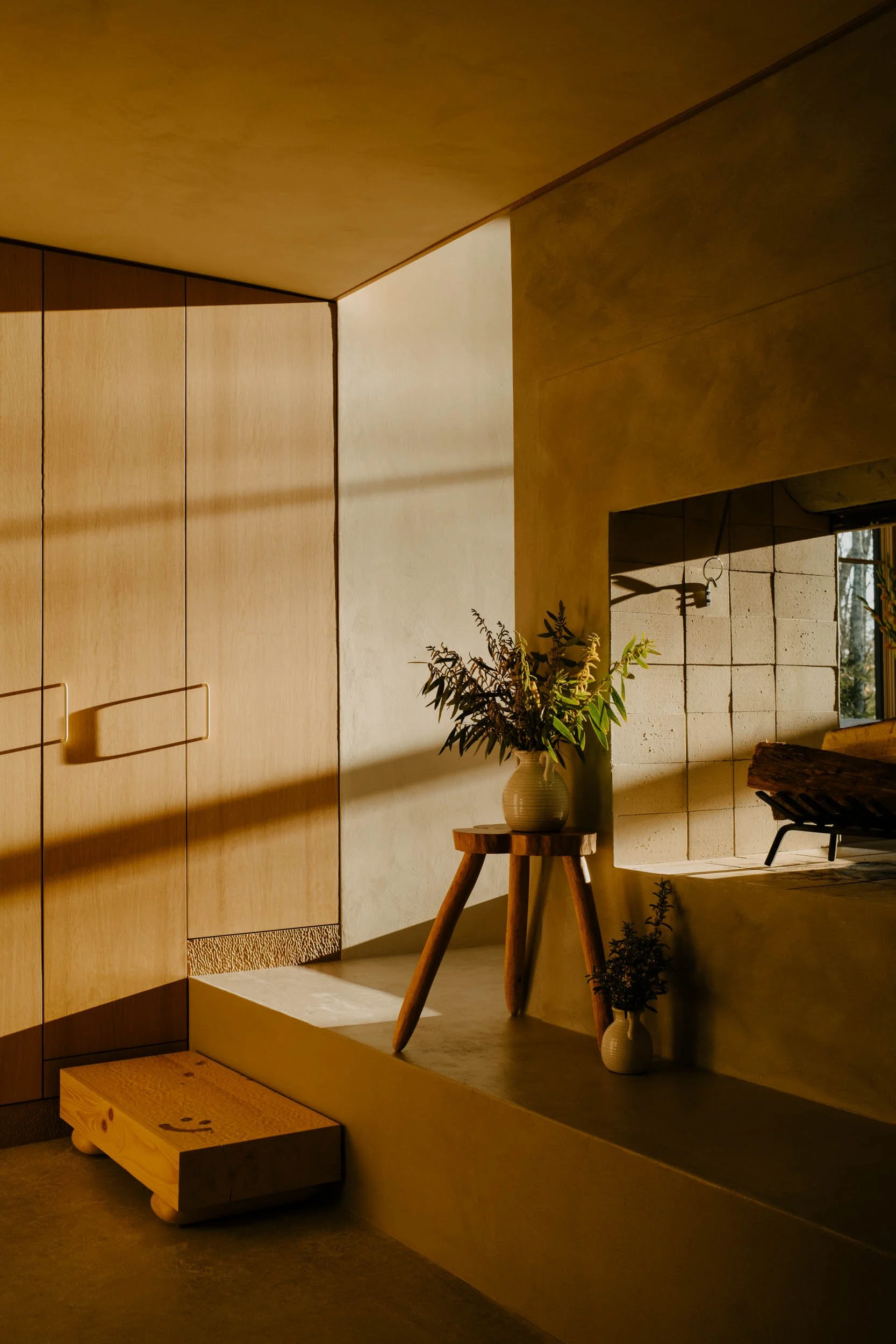 A home entry with wooden cabinets, a small wooden stool, and hand-built fireplace, bathed in warm sunlight.