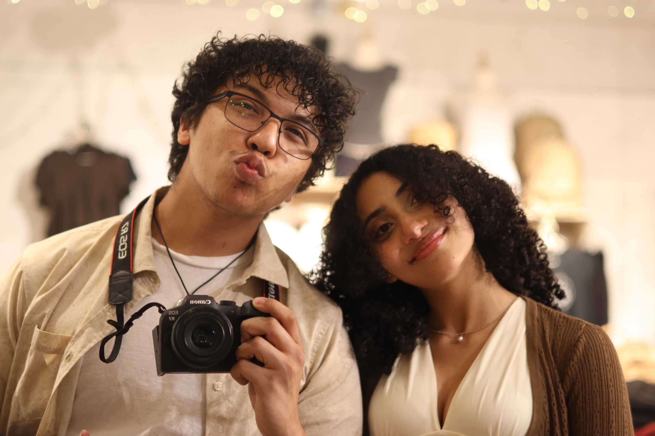 A man with curly dark hair, glasses, and a camera around his neck making a kiss face next to a woman with curly dark hair smiling, both in an indoor setting with blurred warm lighting and background.