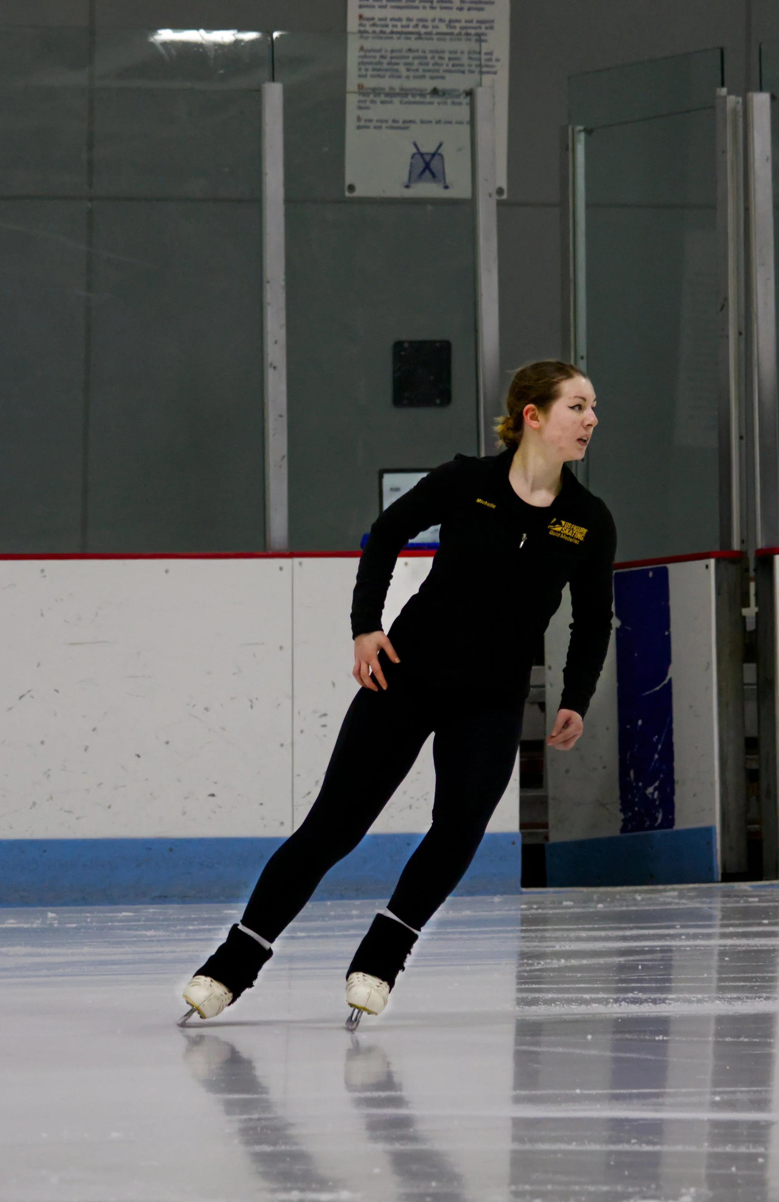 A young woman figure skating on an indoor ice rink, wearing black attire and white ice skates.