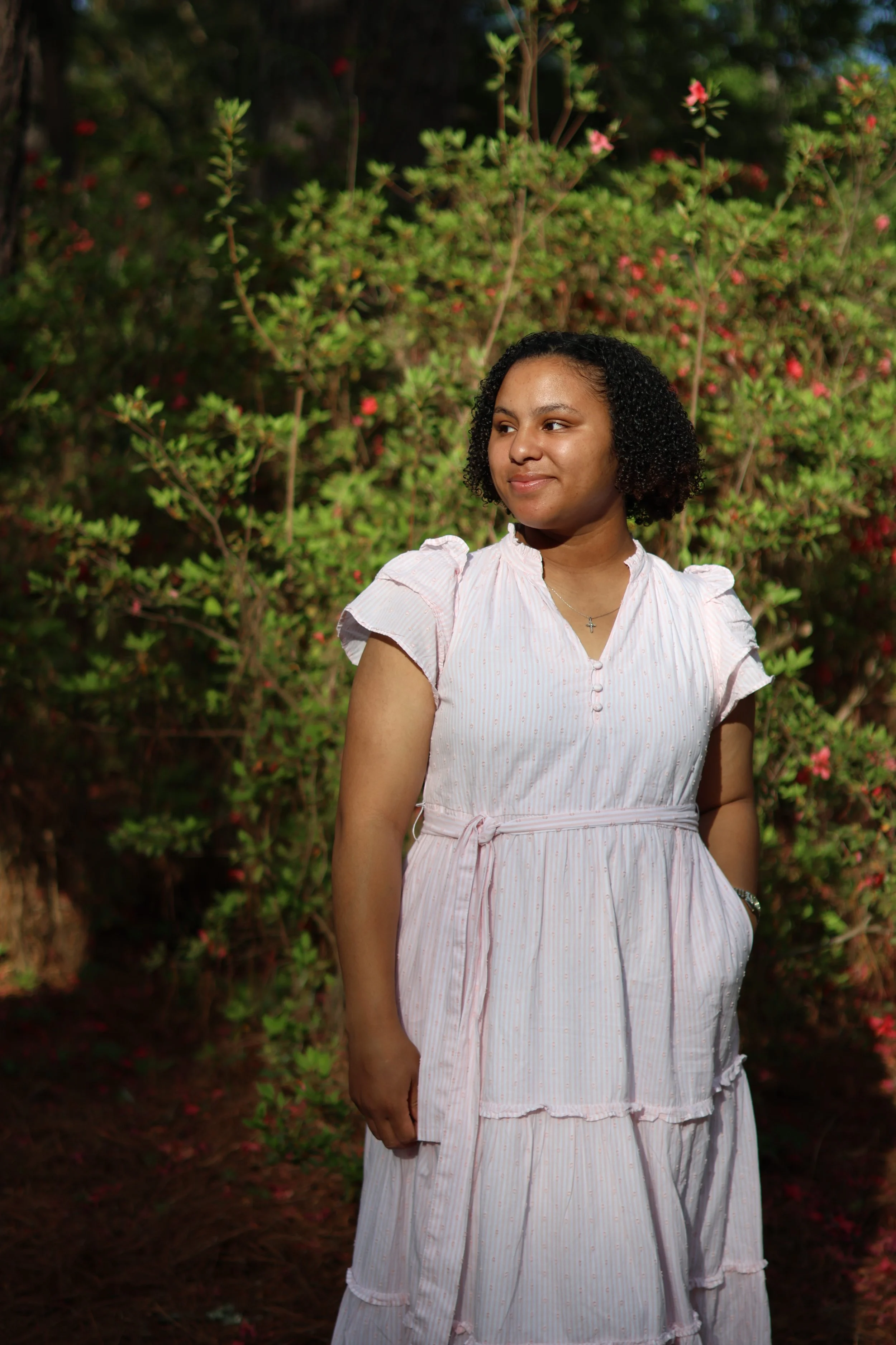 A young woman with curly hair wearing a light pink, short-sleeved dress with a sash standing outdoors in front of bushes with pink flowers, gazing to her right with a slight smile.