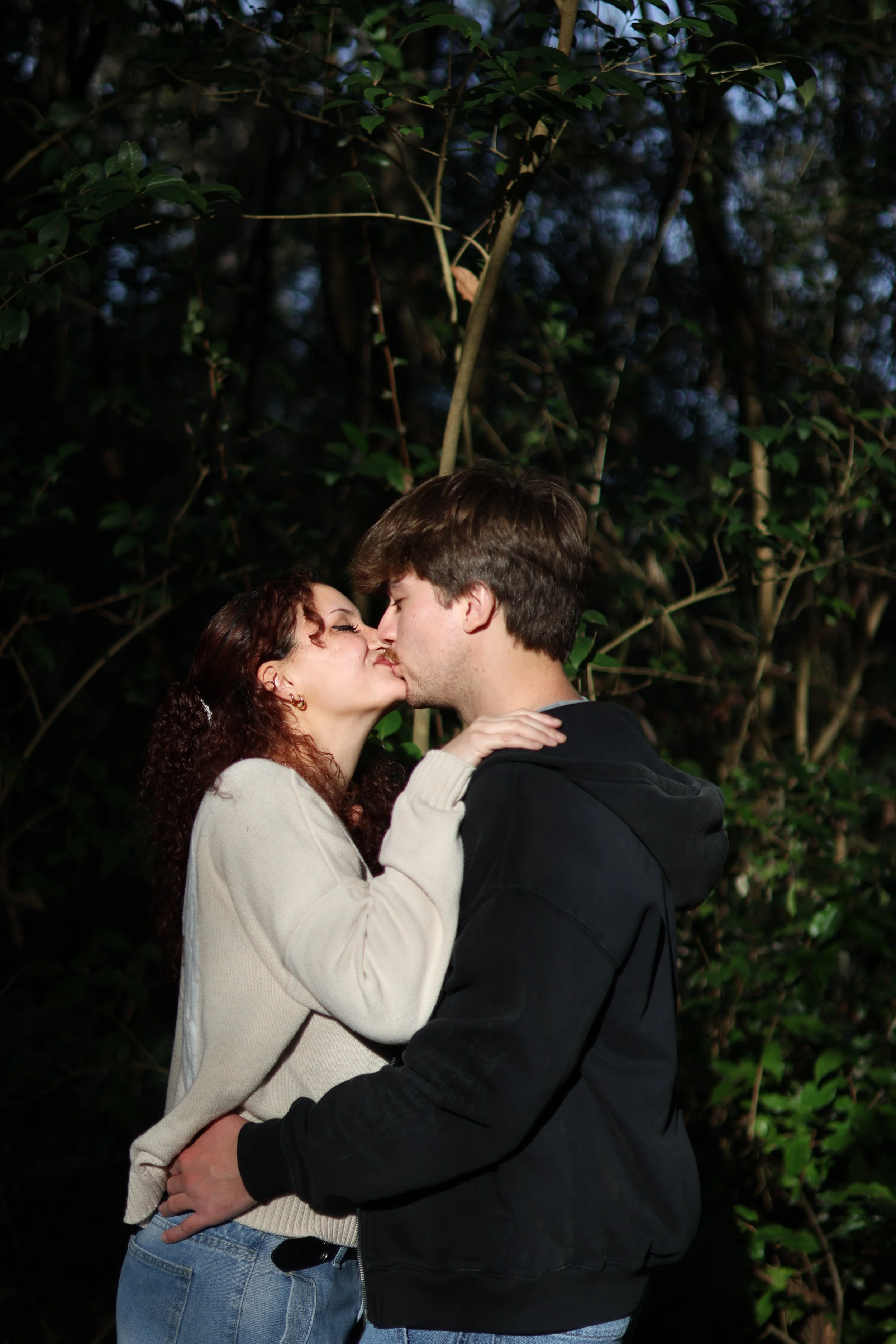 A couple sharing a kiss outdoors at night, surrounded by trees and foliage.