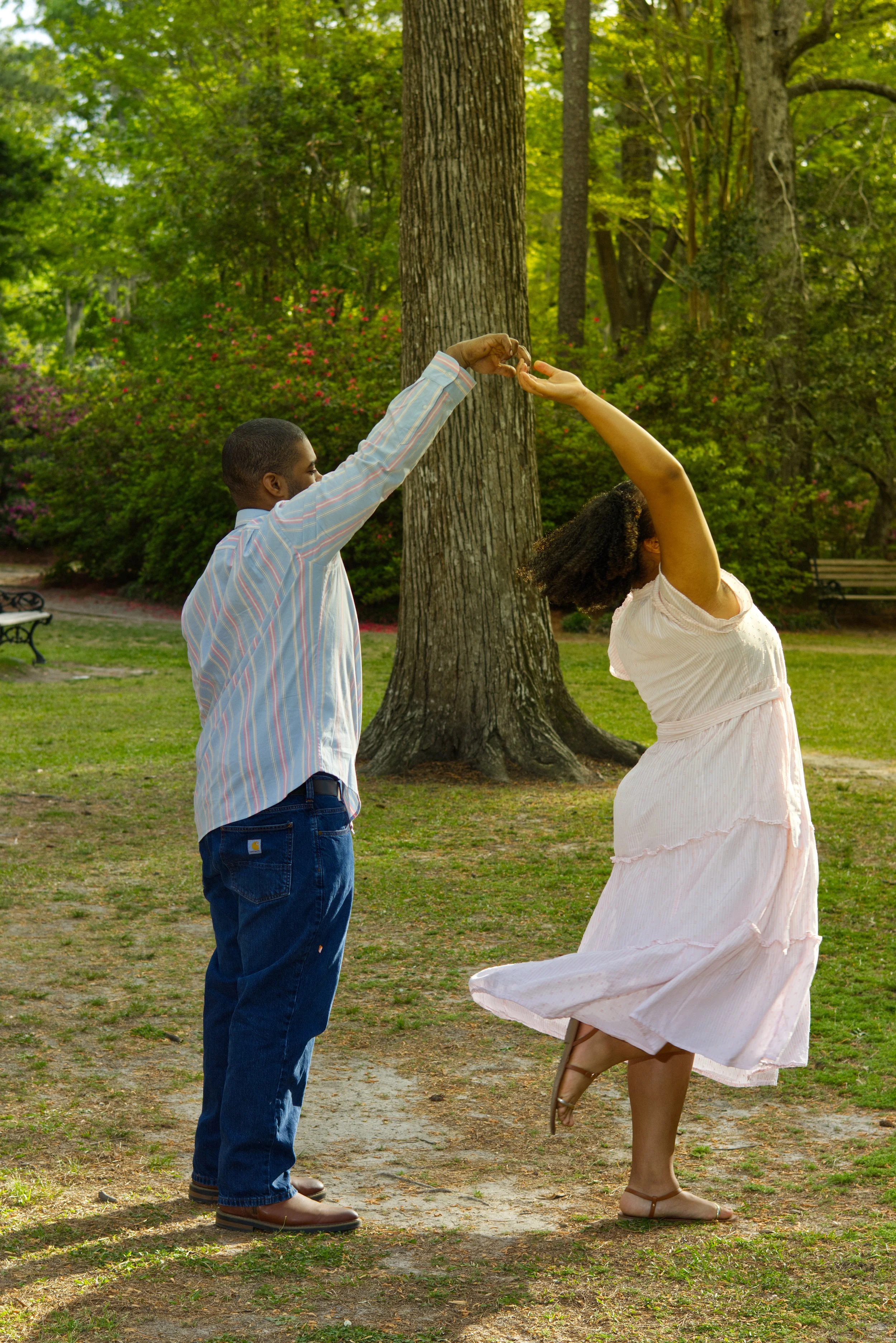 A couple dancing in a park with a large tree in the background, green foliage, and benches.
