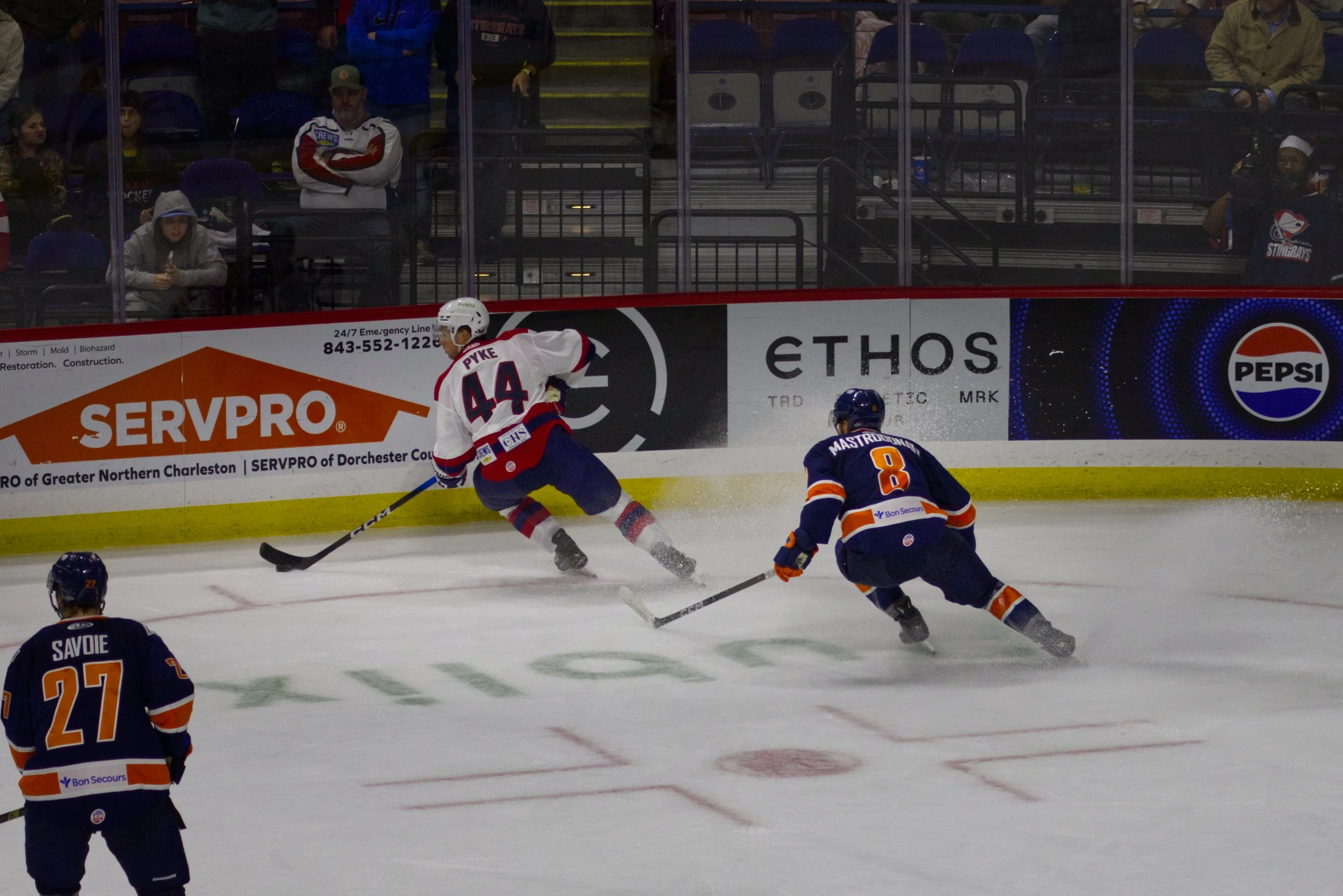 Ice hockey game with players in blue and white jerseys, one skater in white jersey number 44 and another in blue jersey number 8, pursuing the puck near the boards. Several spectators watch from the stands.