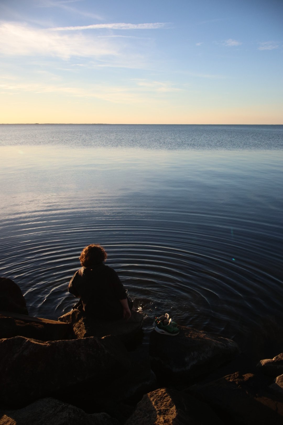 Person sitting on rocks by the water during sunset, with calm water and a distant horizon under a partly cloudy sky.