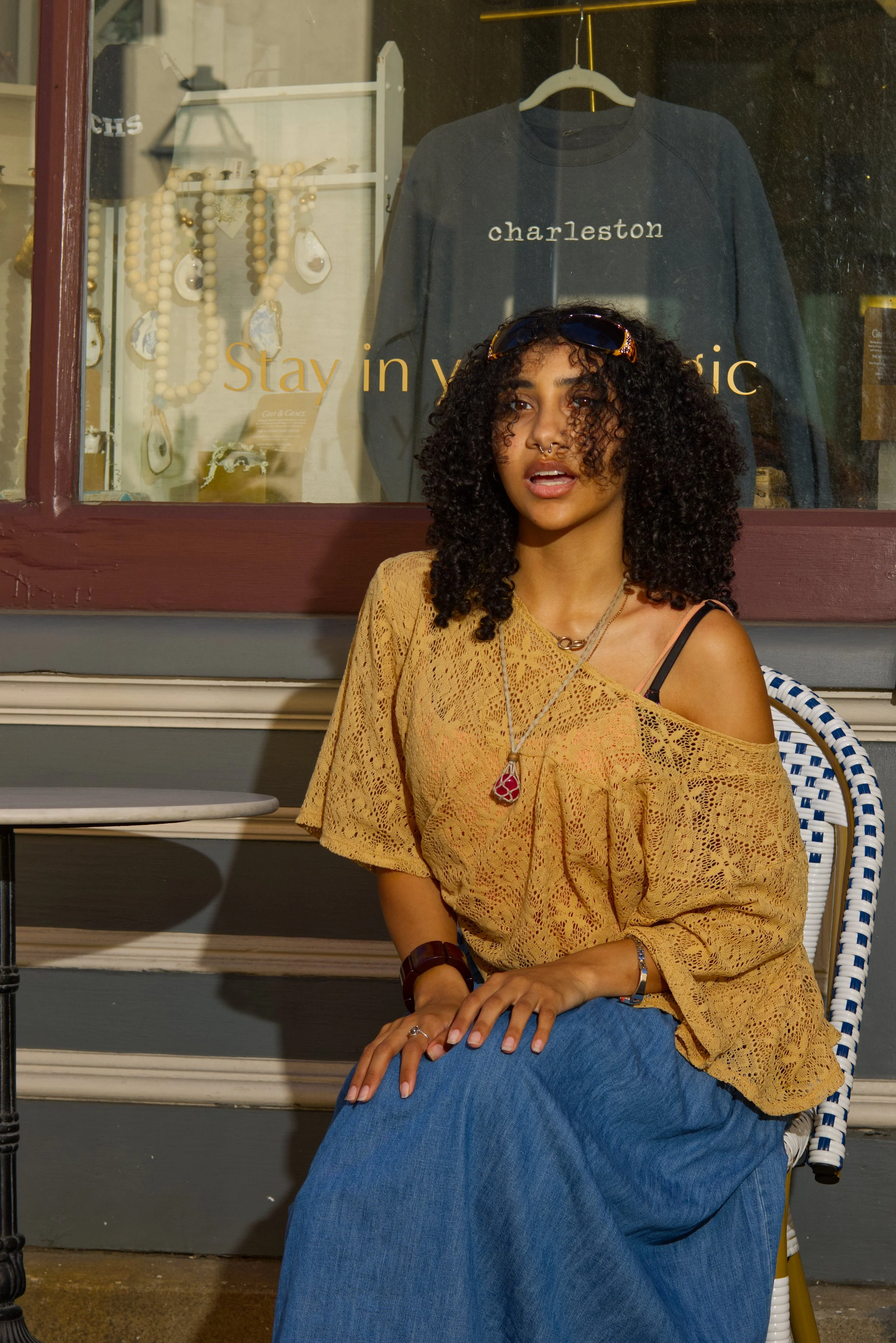 A woman with curly hair, wearing sunglasses on her head, a yellow lace off-shoulder top, and a long blue skirt, sitting outside a store with jewelry and clothing in the window.