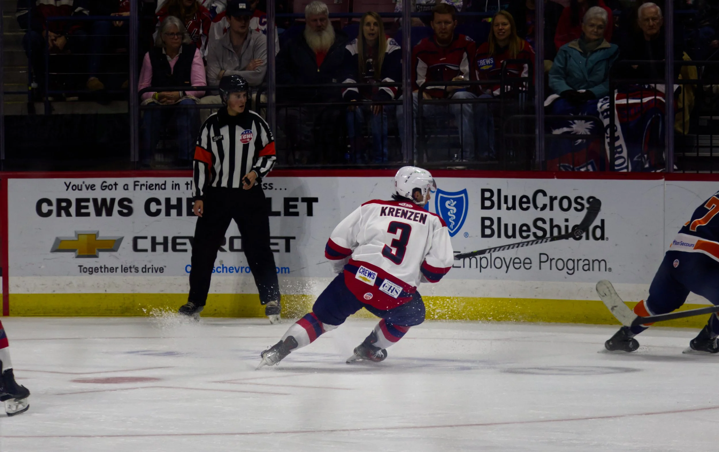 Ice hockey game with a player in a white jersey number 3, skating on the ice near the boards, with spectators watching behind the glass. An official referee is standing nearby.