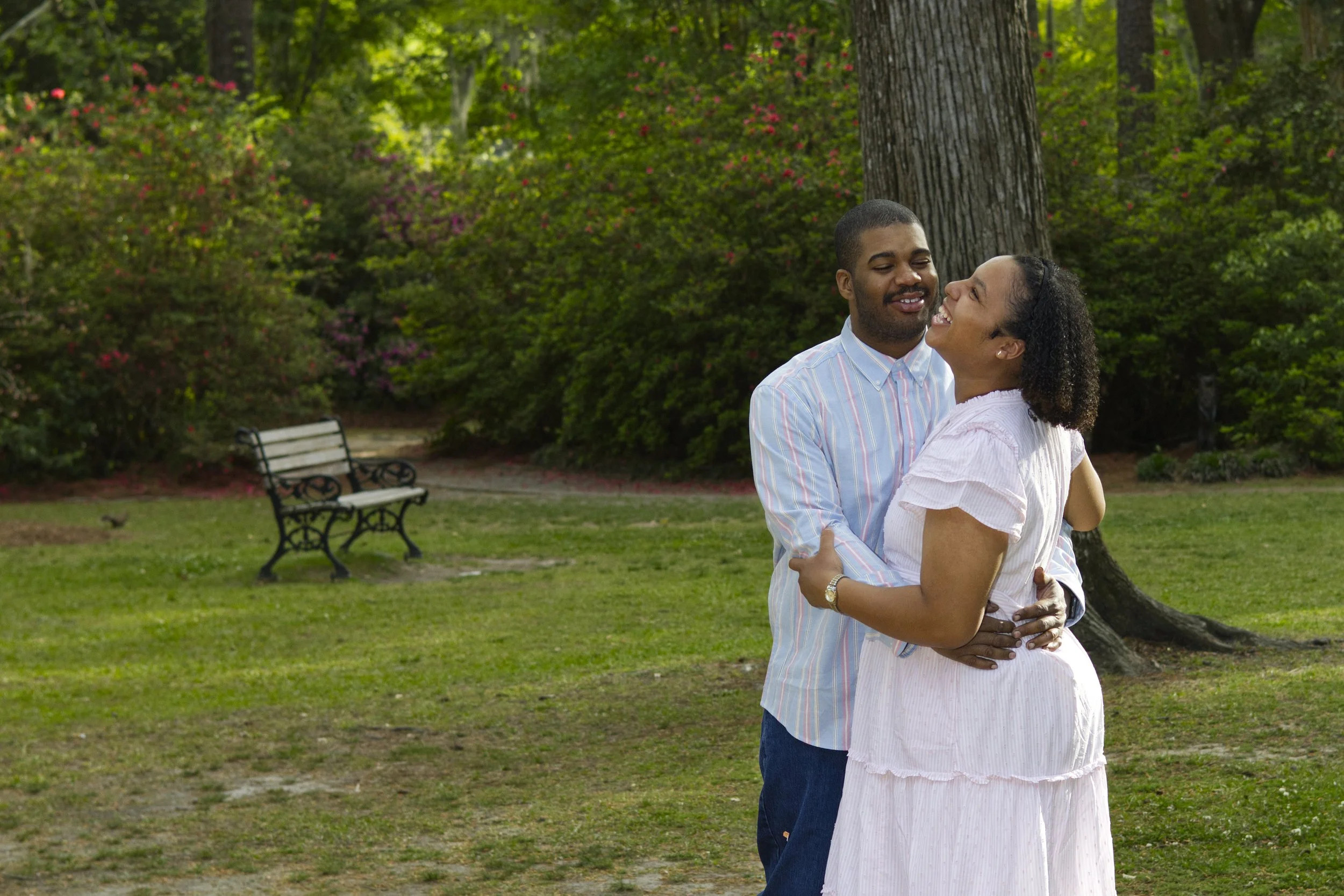 A couple embracing and smiling at each other in a park with trees, shrubs, and a bench in the background.