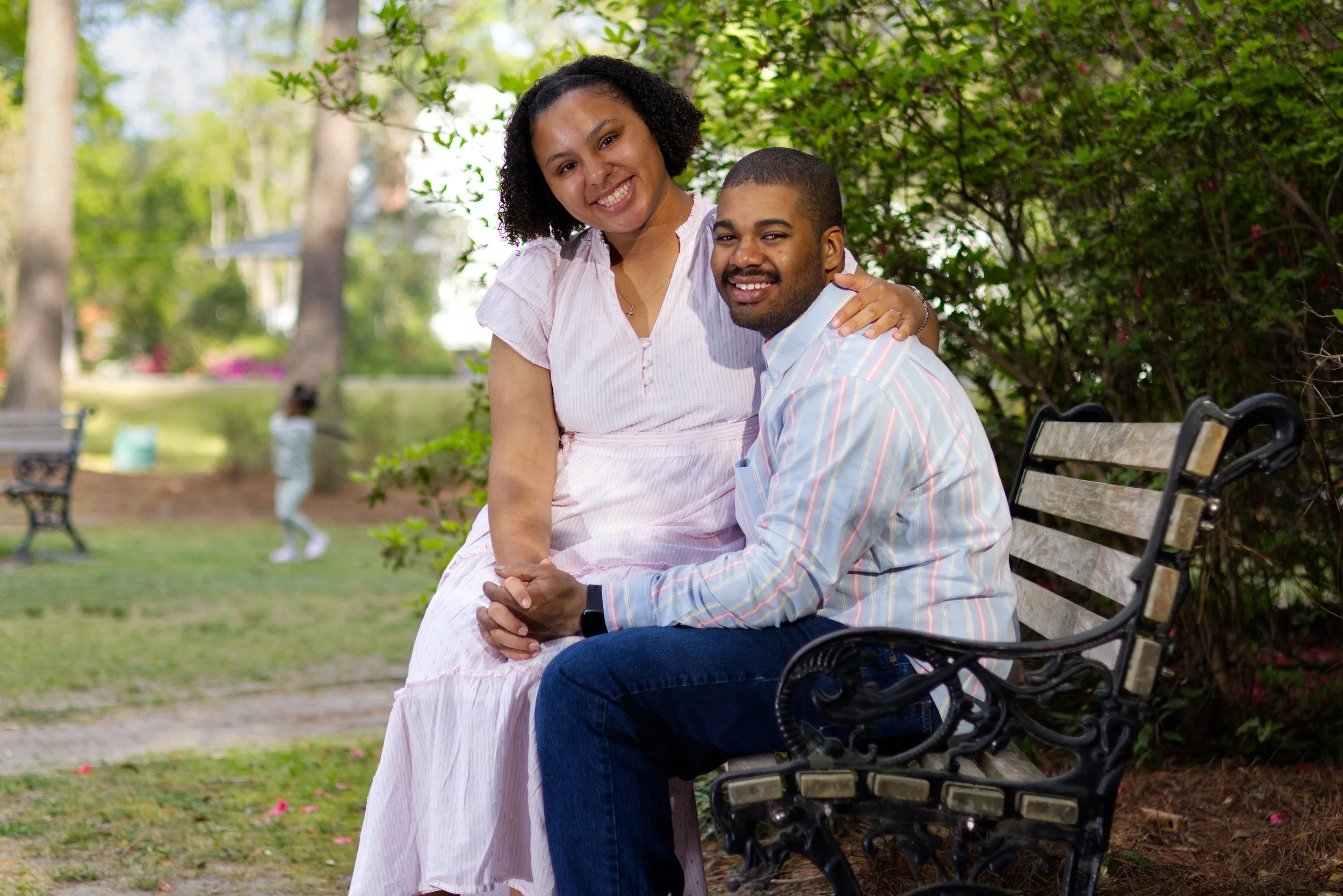 A smiling couple sitting on a park bench, with the woman sitting on the man's lap, surrounded by green trees and a blurry child playing in the background.